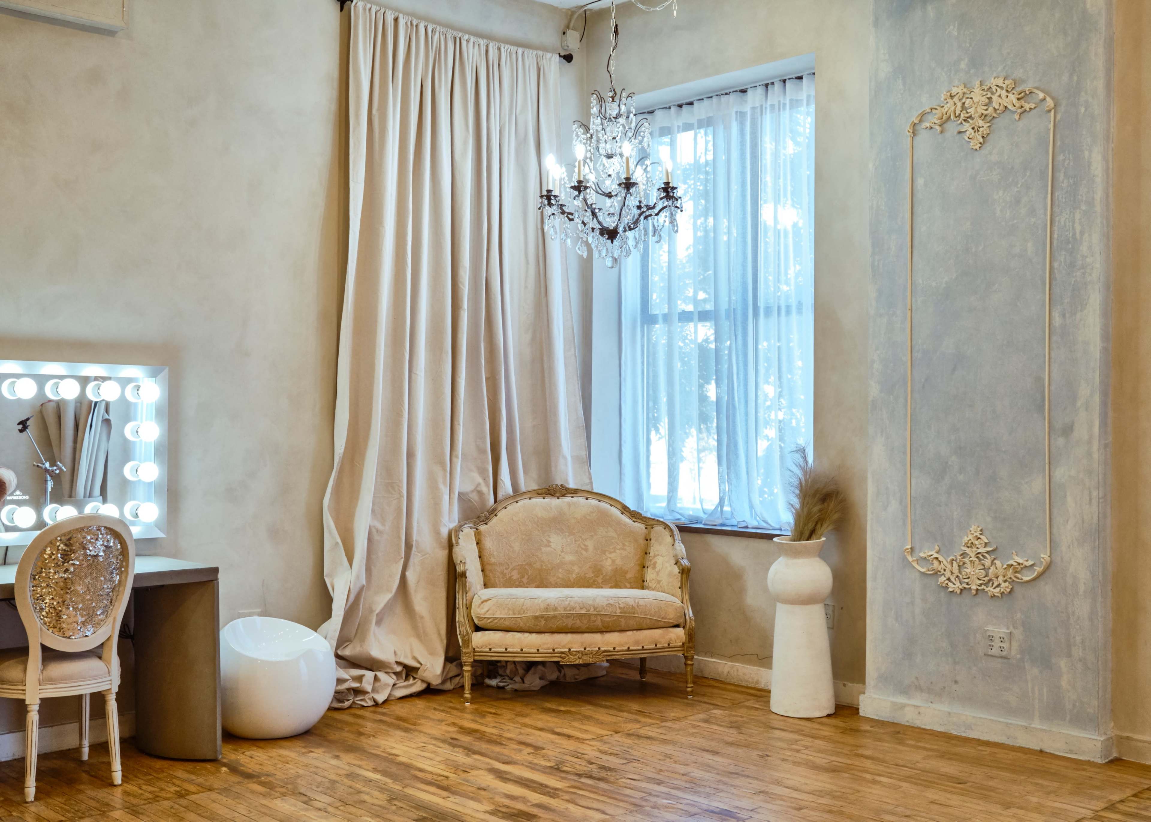 The image shows a room with a vintage-style couch, a modern vanity with lights, and a large window adorned with sheer curtains.