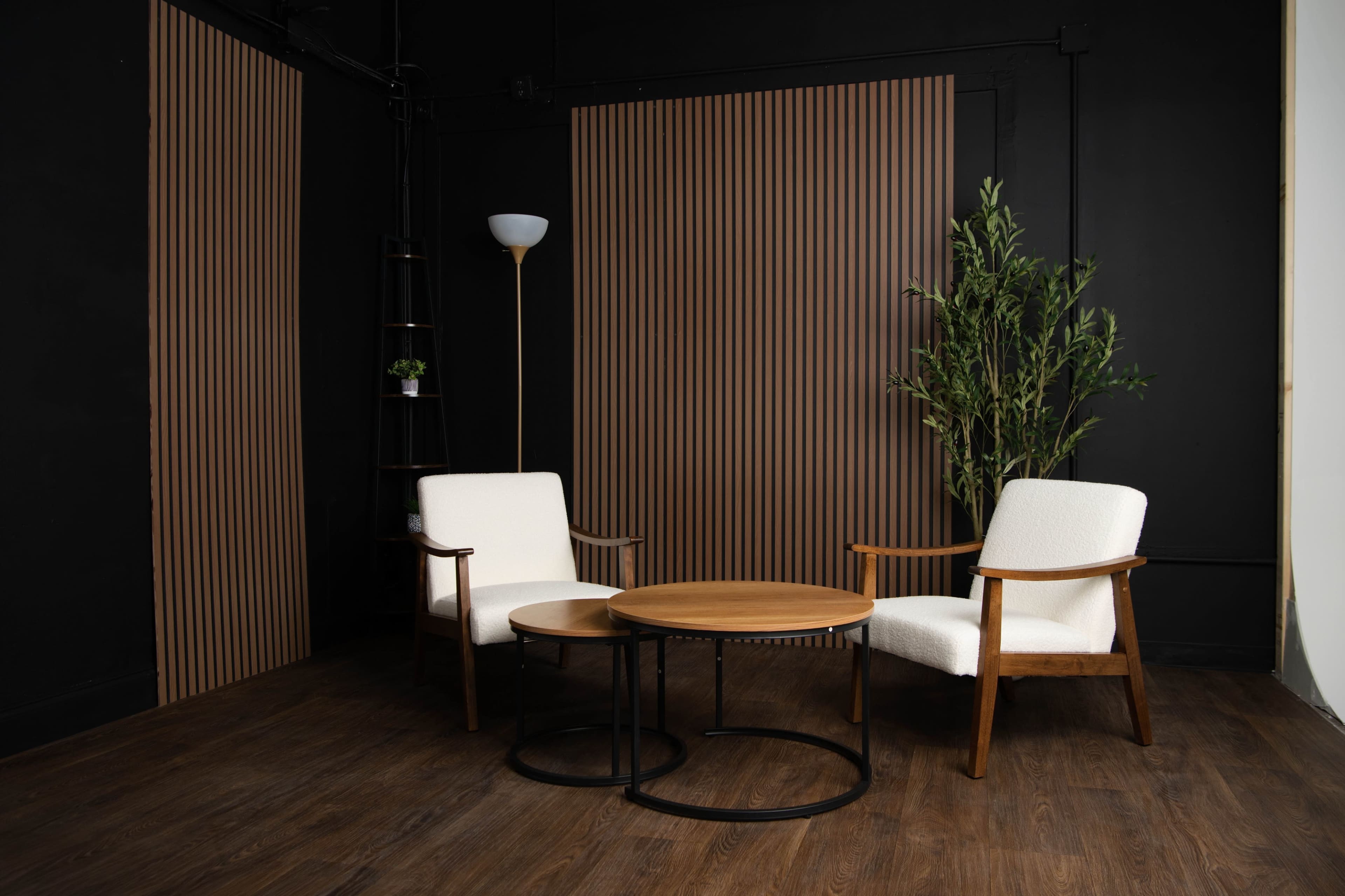 The image depicts a minimalist interior with two white armchairs, a round wooden table, and a potted plant, set against dark walls with vertical wooden panels.