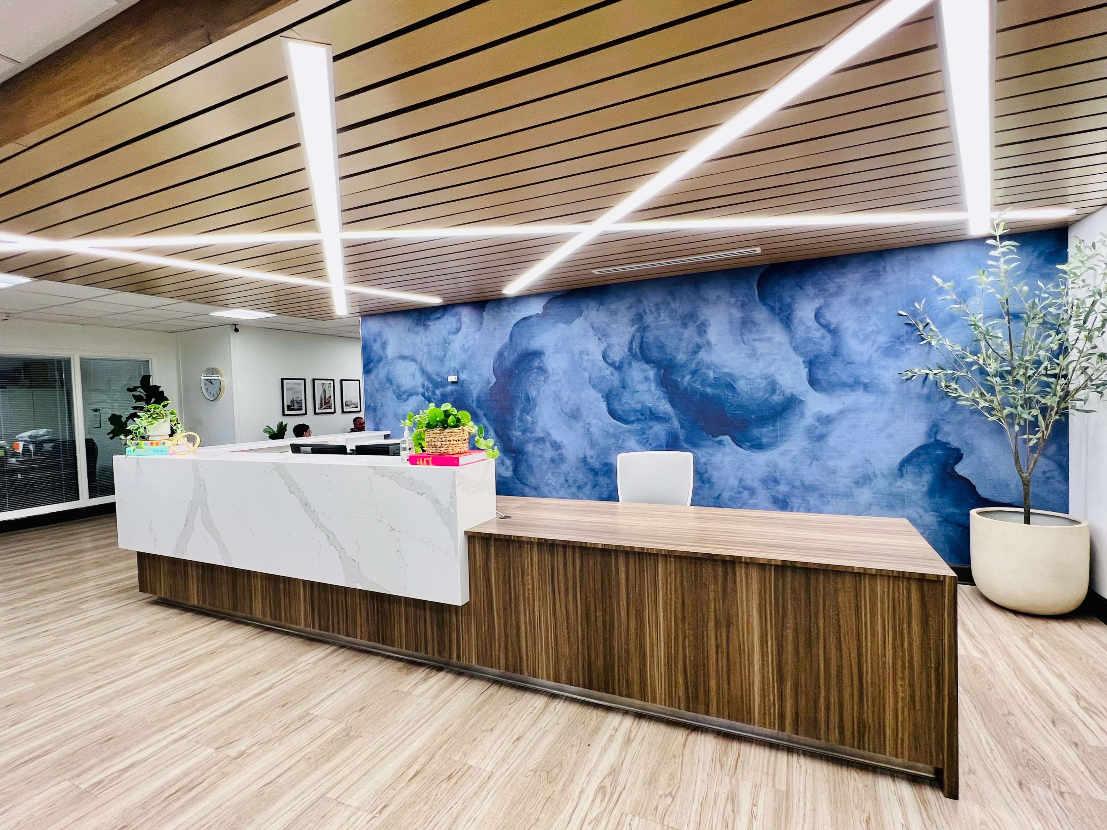 A modern reception area featuring a white marble front desk, a wooden countertop, and a blue textured wall with overhead lighting.