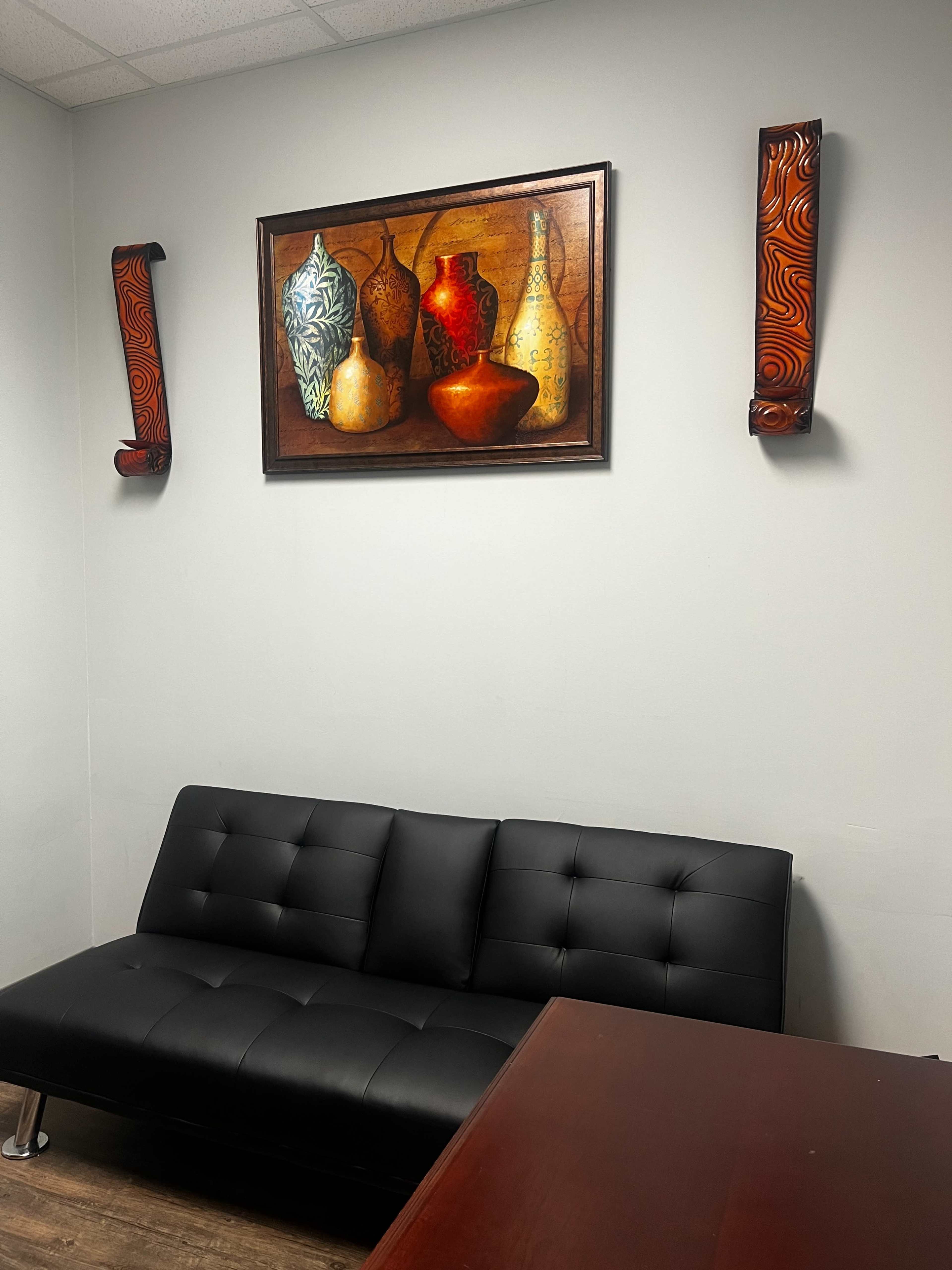 A small office space with a black futon, a wooden table, and abstract artwork featuring vases hanging on the walls.