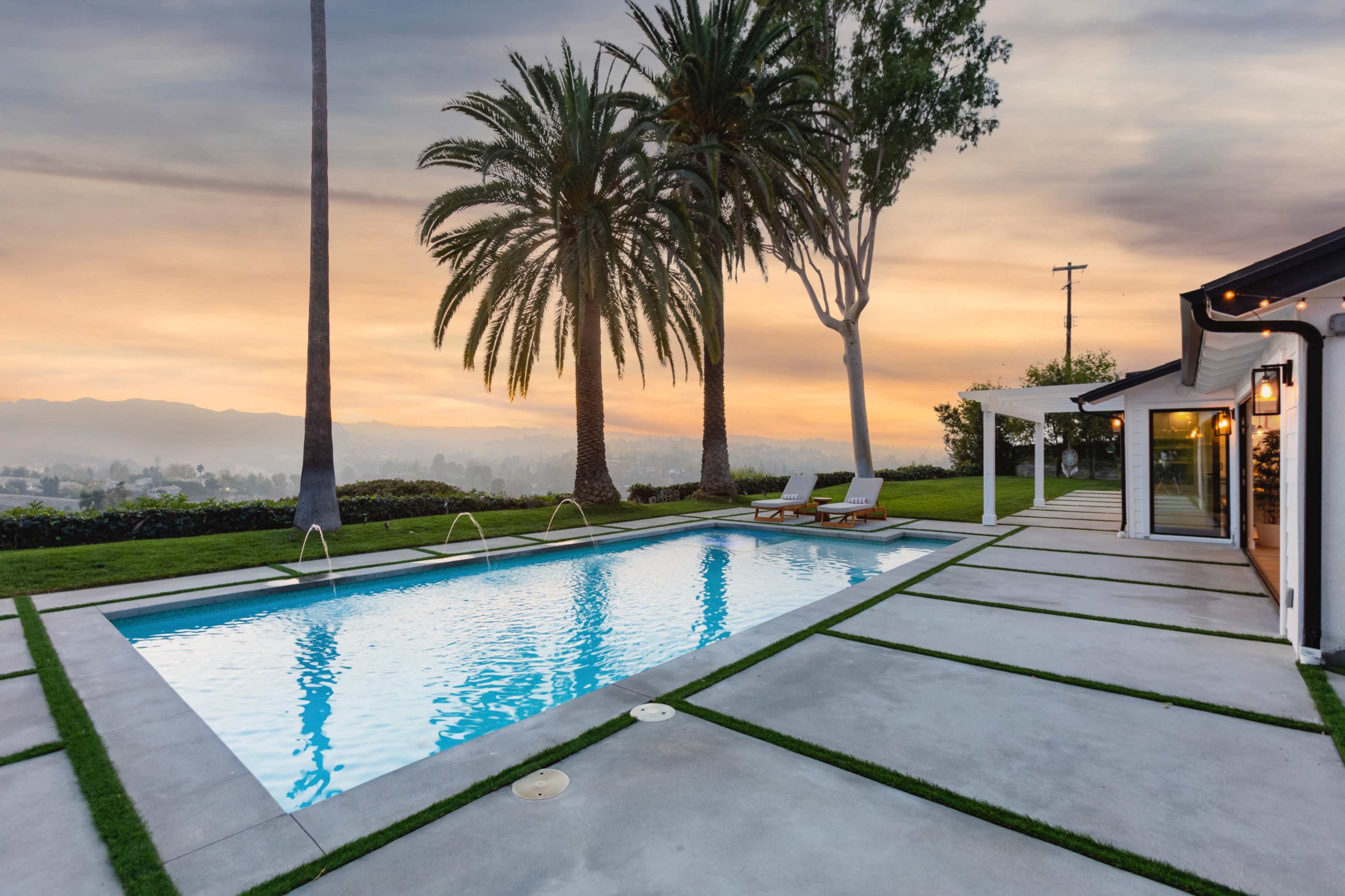 A swimming pool surrounded by palm trees and a concrete deck overlooks a scenic sunset view.