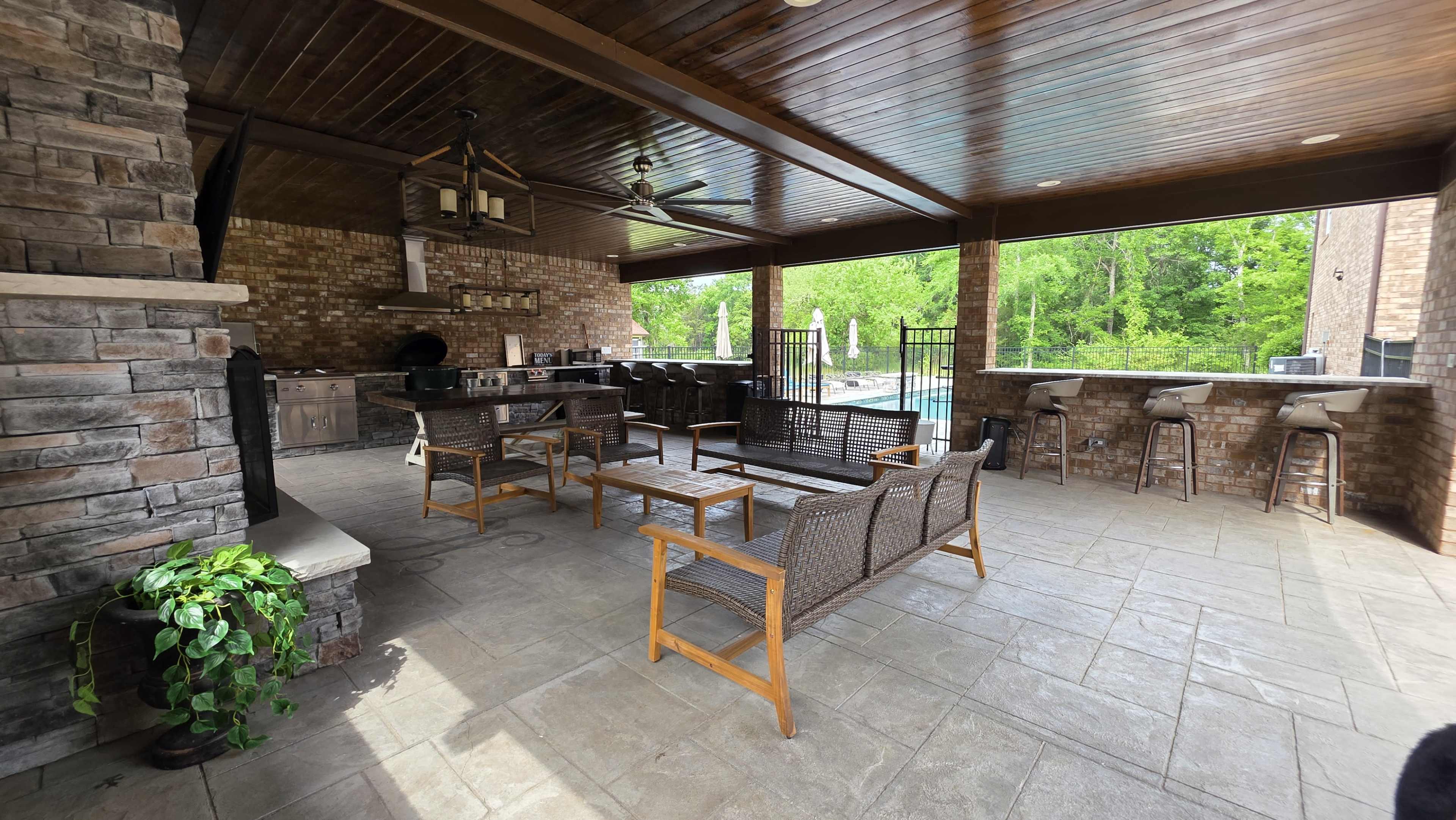 The image shows a covered outdoor area featuring a stone barbecue grill, several wooden seating arrangements, and a swimming pool visible beyond the space surrounded by greenery.