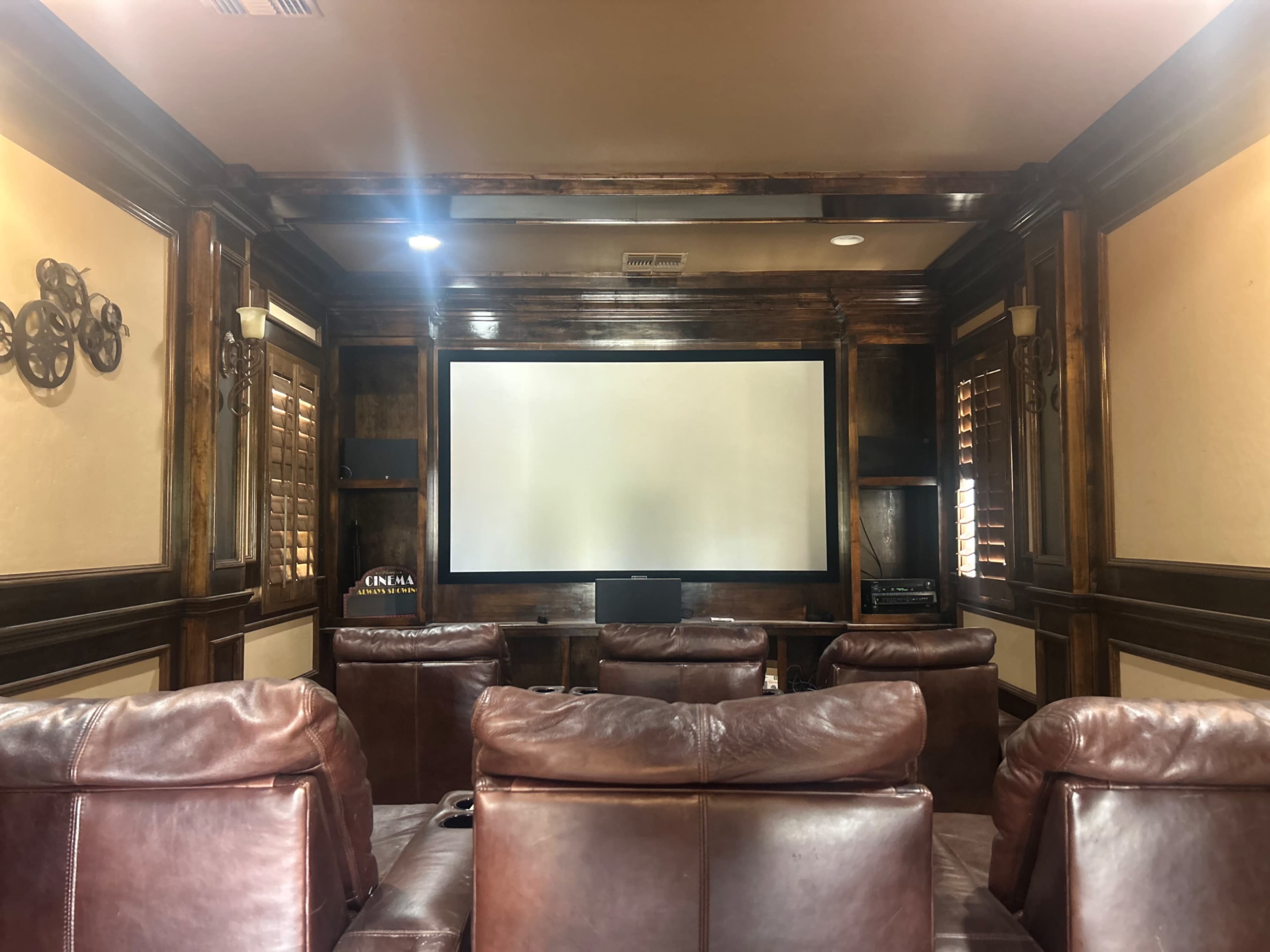 The image depicts a home theater with brown leather recliners facing a large, blank screen framed by wooden paneling.