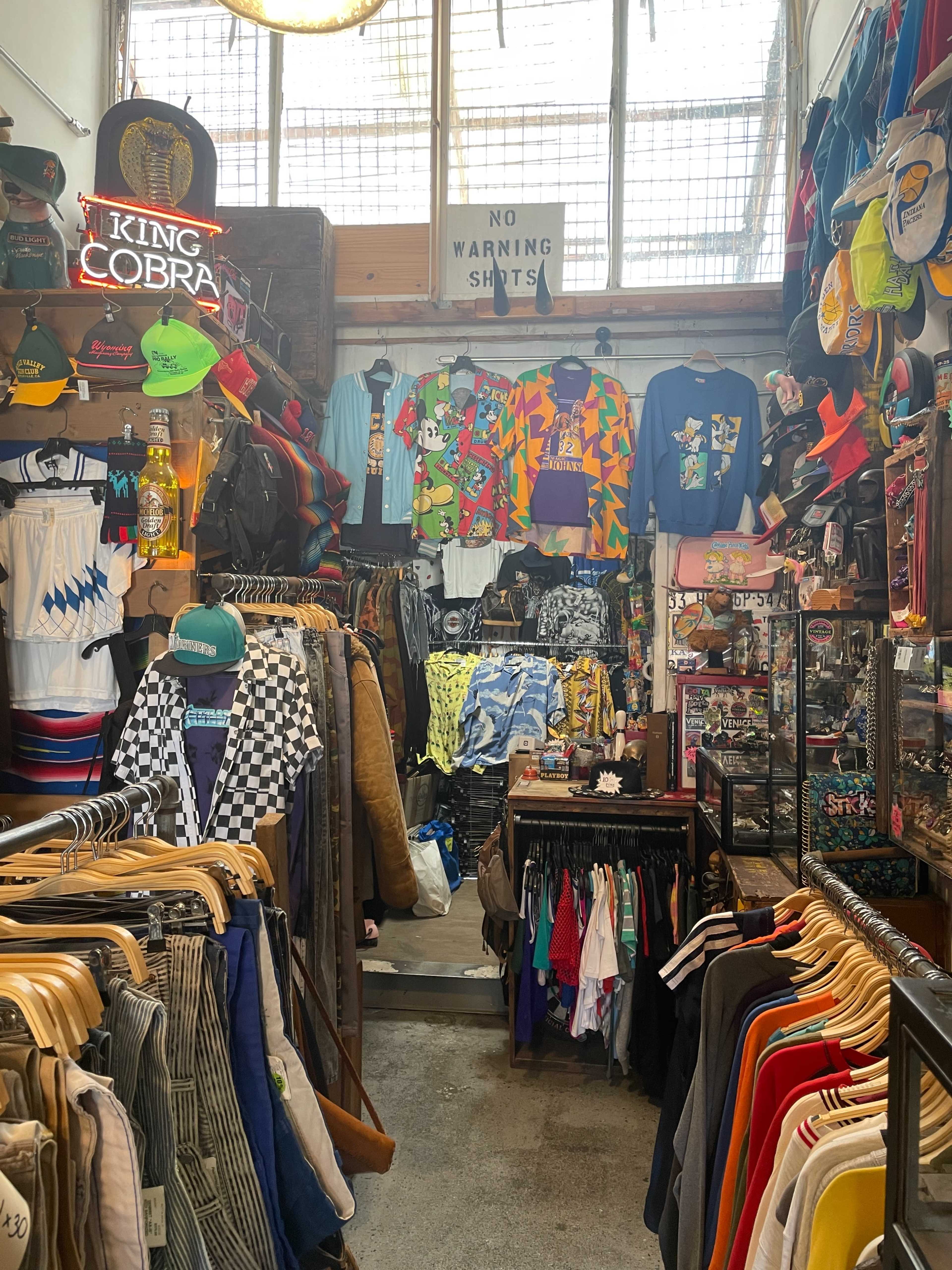 The image shows a vintage clothing store filled with racks of colorful apparel and various accessories displayed on walls.