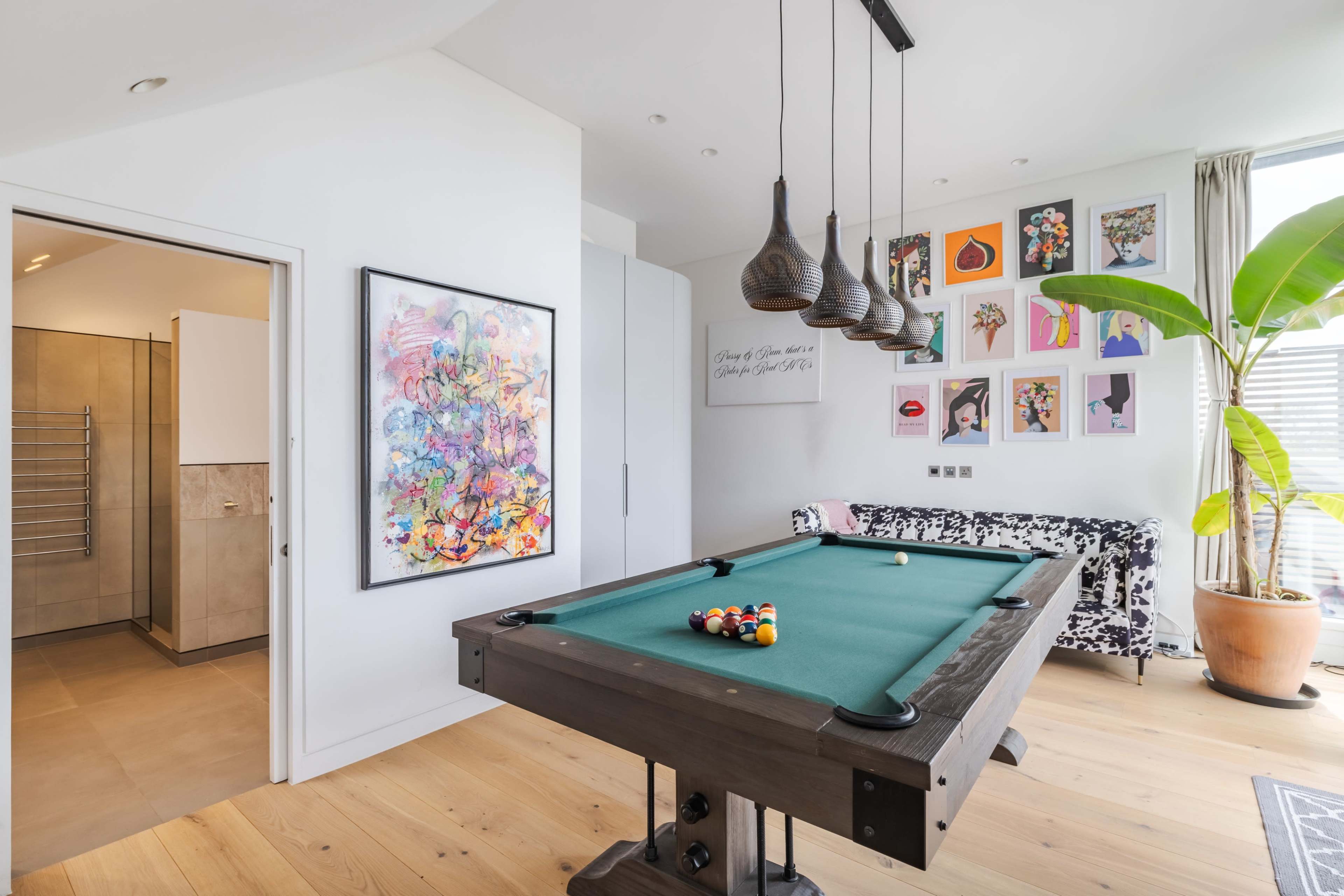 A billiard table is positioned in a well-lit room that features an art piece, a decorative sofa, and a plant, with an open doorway leading to a bathroom area.
