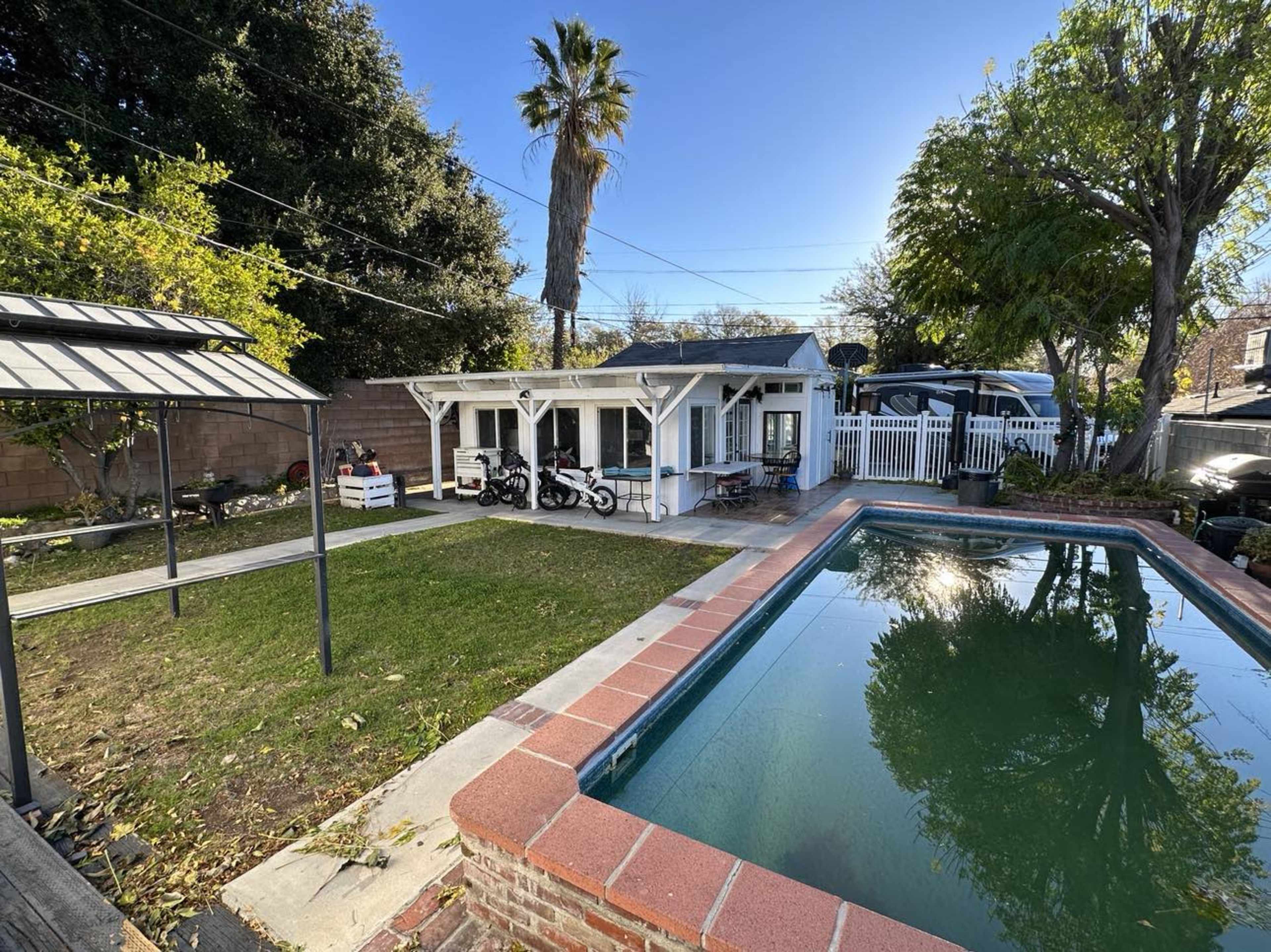 The image shows a backyard with a swimming pool, lawn, a small white building, and two motorcycles parked nearby.