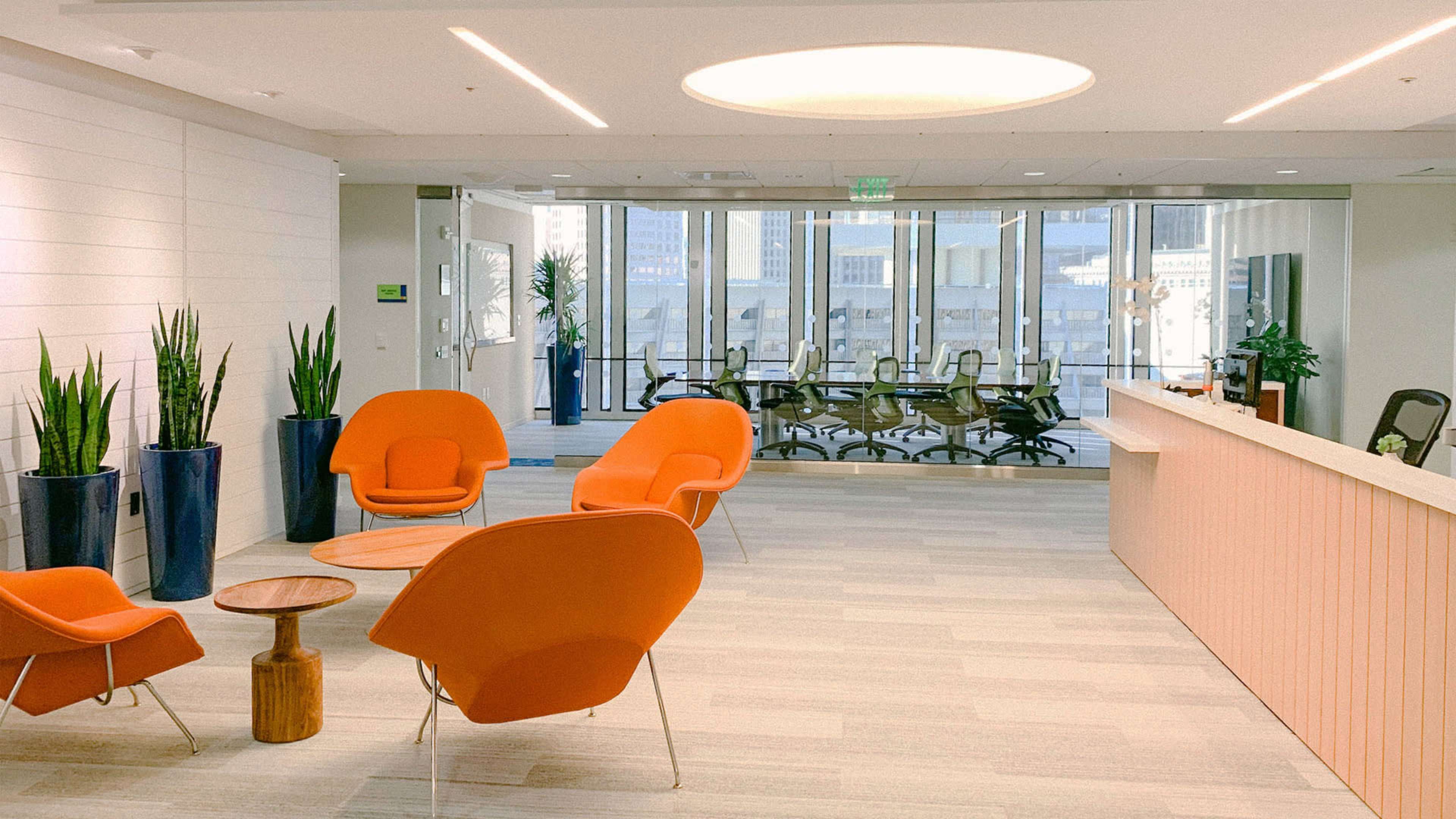The image shows a modern office reception area with orange chairs, a wooden table, and a conference room visible through large windows.