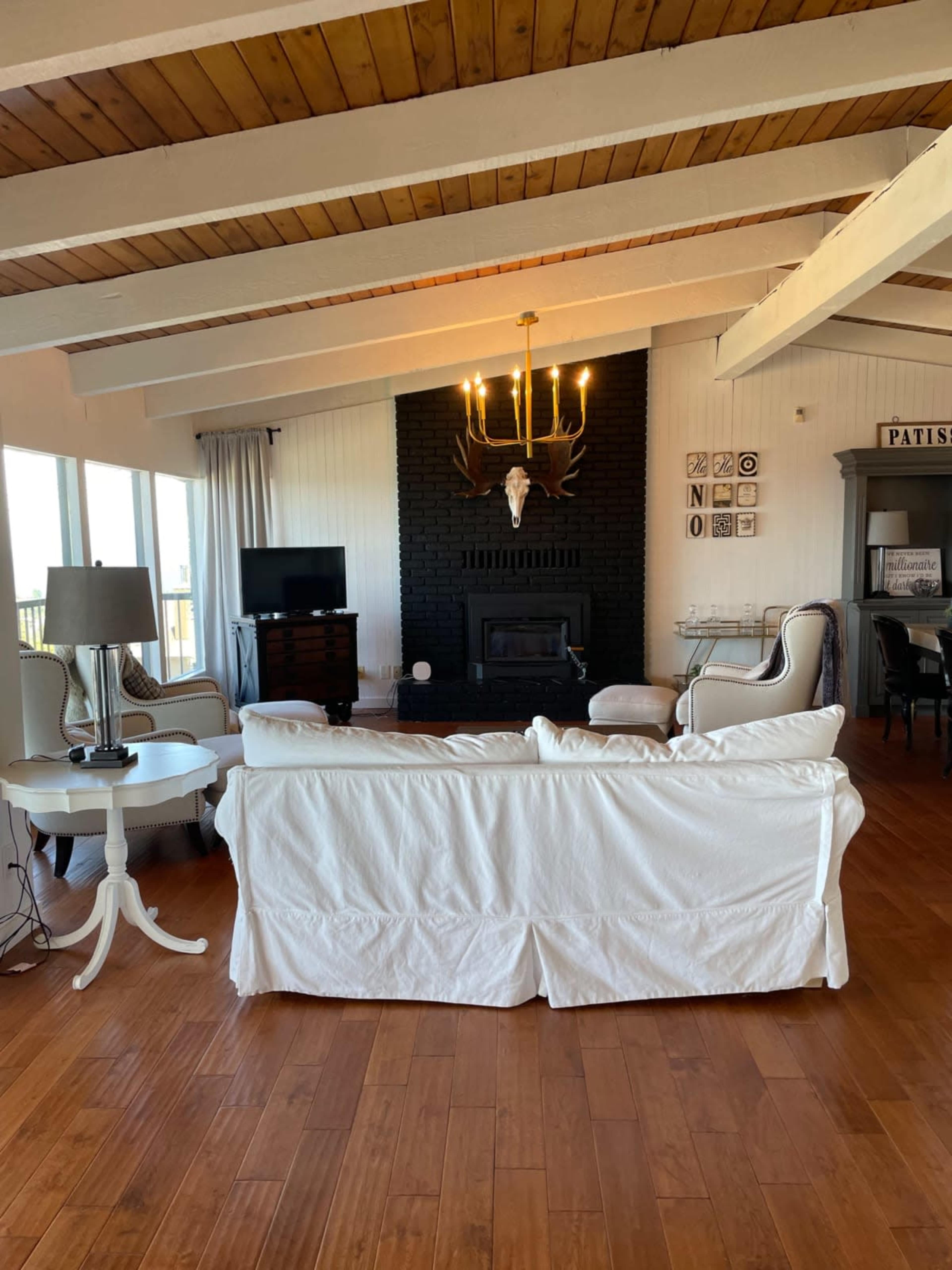 A living room features a white couch, two armchairs, a black fireplace with a mounted deer head, and a television against a wall of windows.