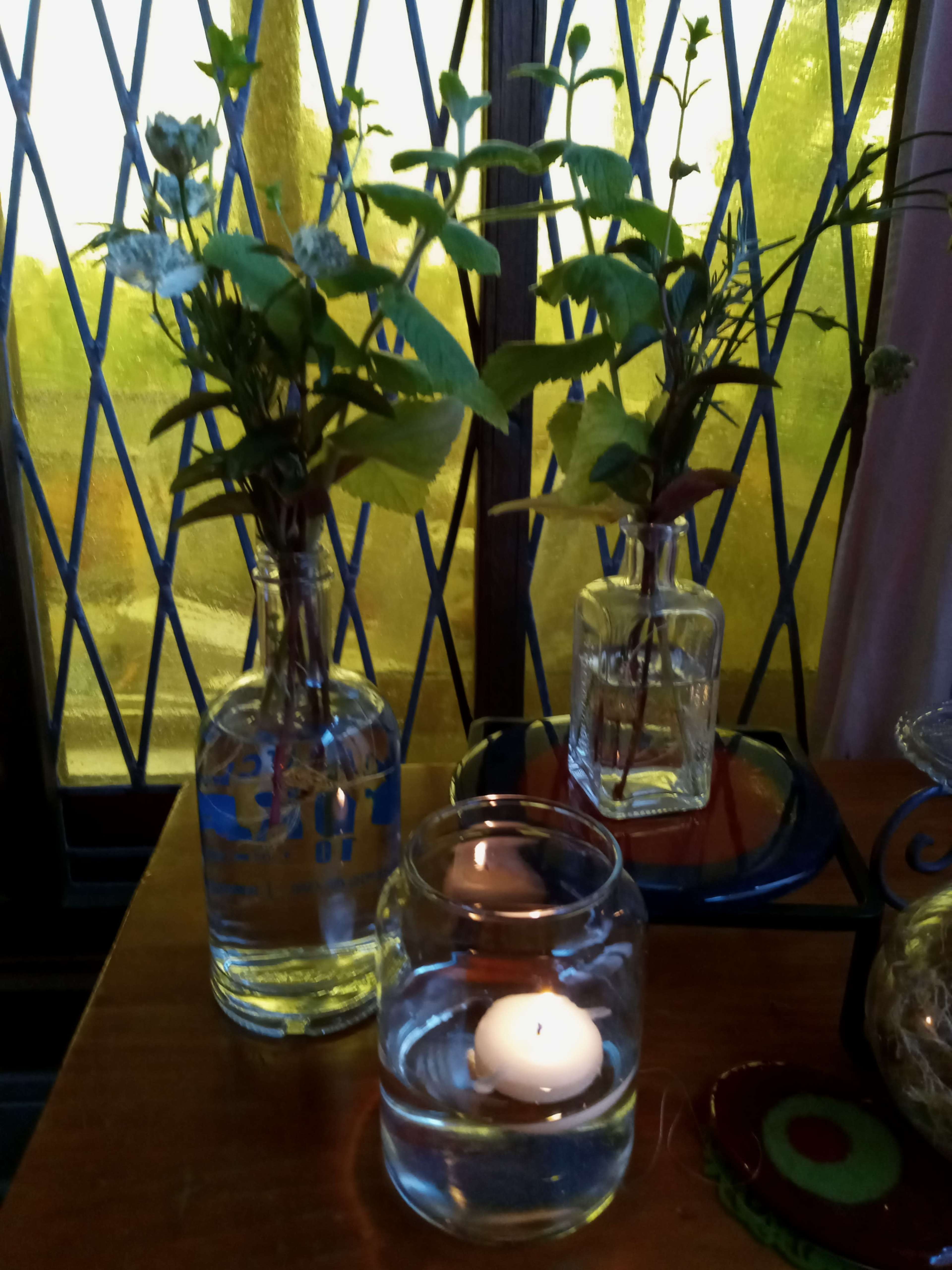 Two vases with greenery and a candle float in a glass jar, situated on a table near a window with stained glass.