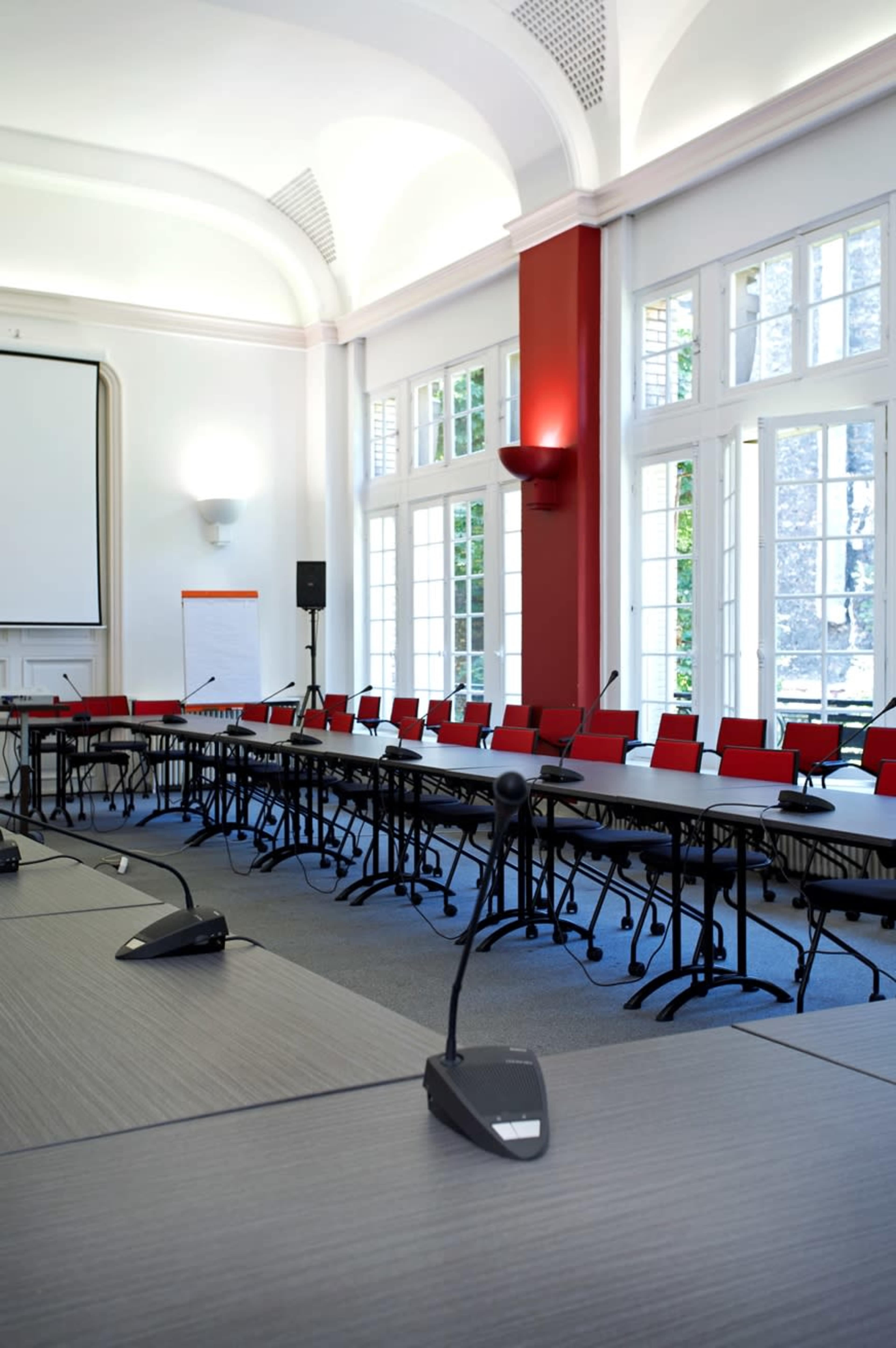 A spacious conference room features a large table with several microphones and rows of red chairs arranged for a meeting.