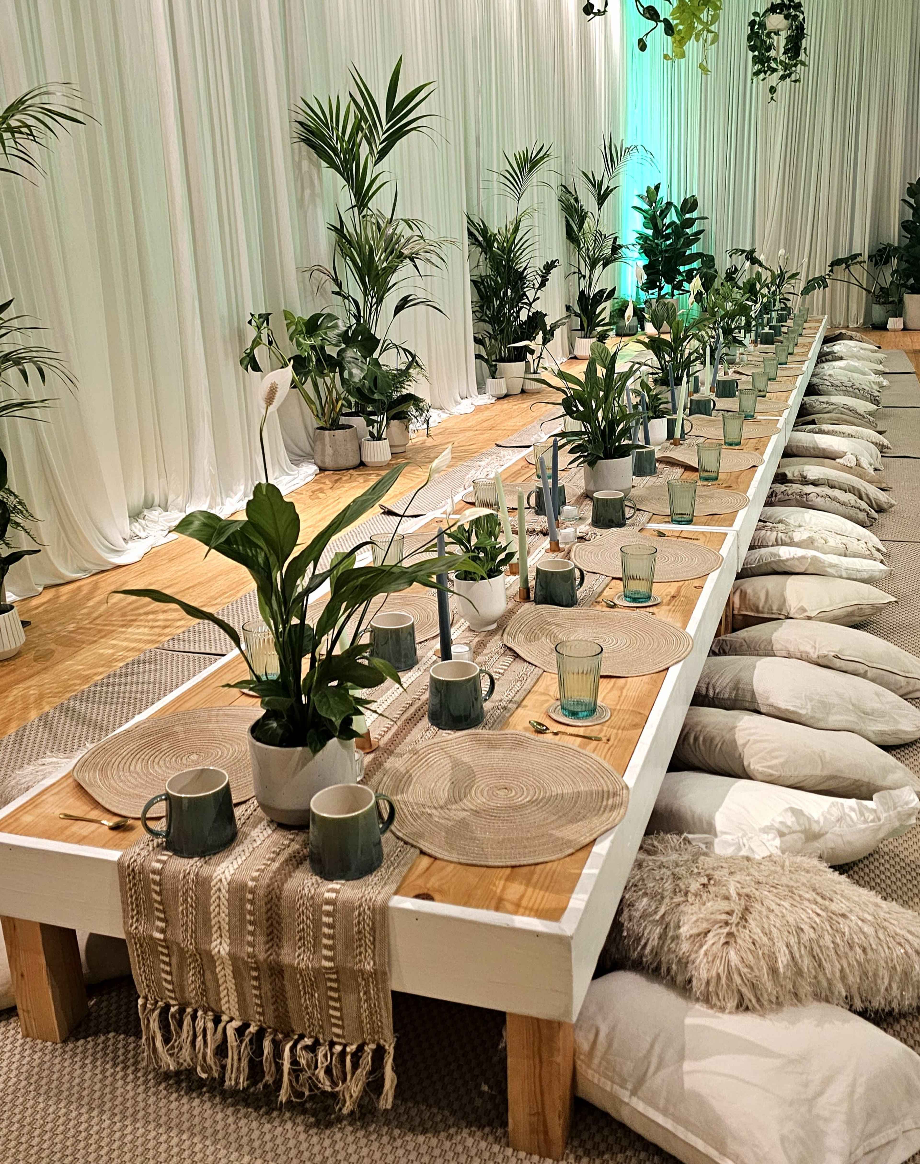 A long wooden table is set with plants, placemats, and cups, surrounded by cushions on the floor and draped with white fabric in a well-lit room.