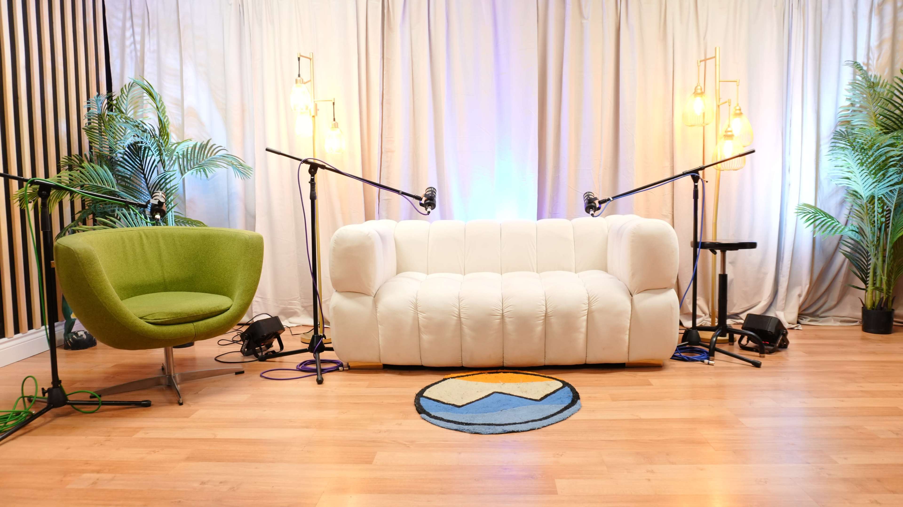 The image features a stylish studio setup with a white couch, a green armchair, and two microphone stands, all set against a backdrop of curtains and decorative plants.