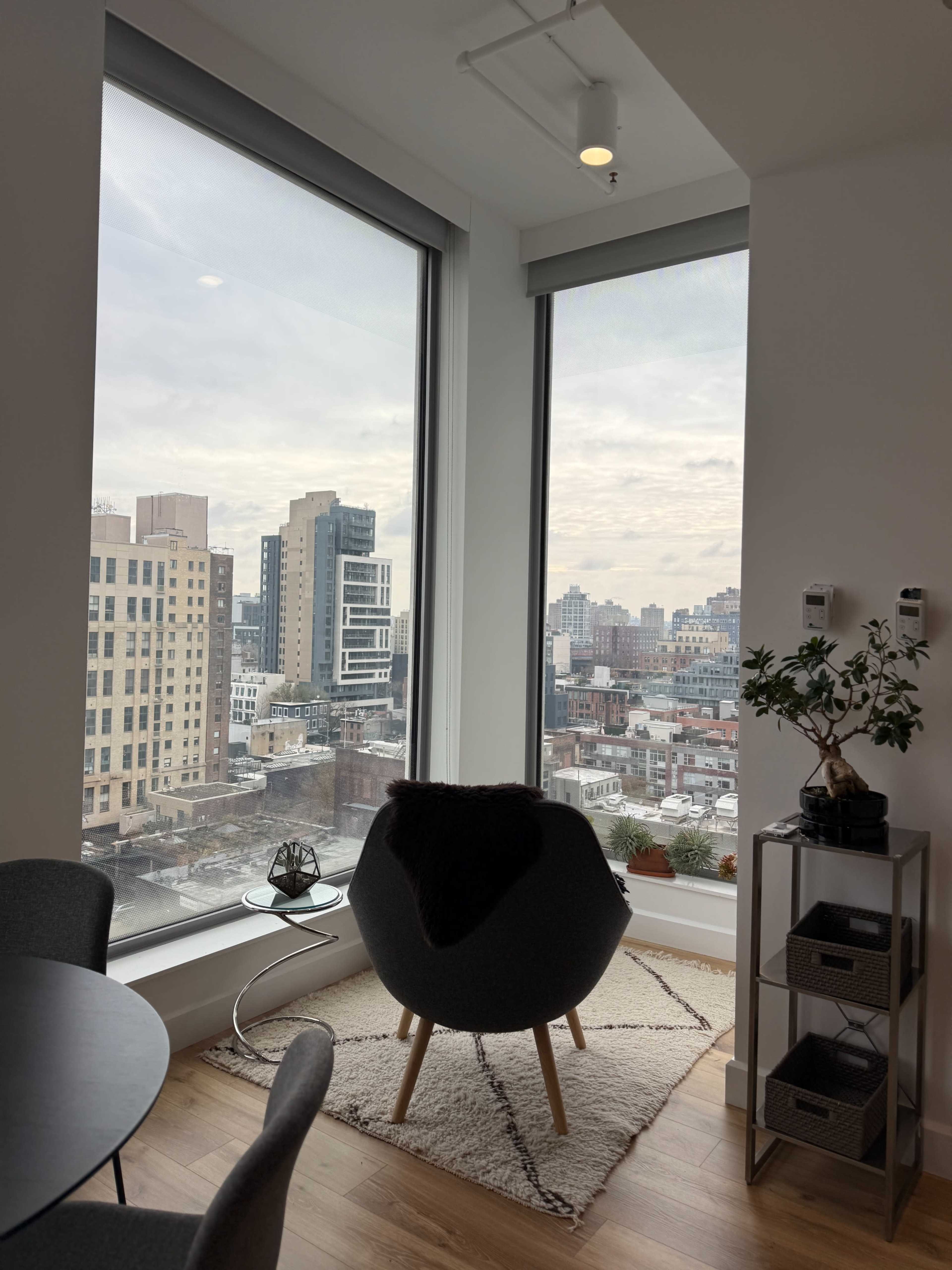 A cozy corner of a modern apartment features a chair next to large windows overlooking a city skyline.