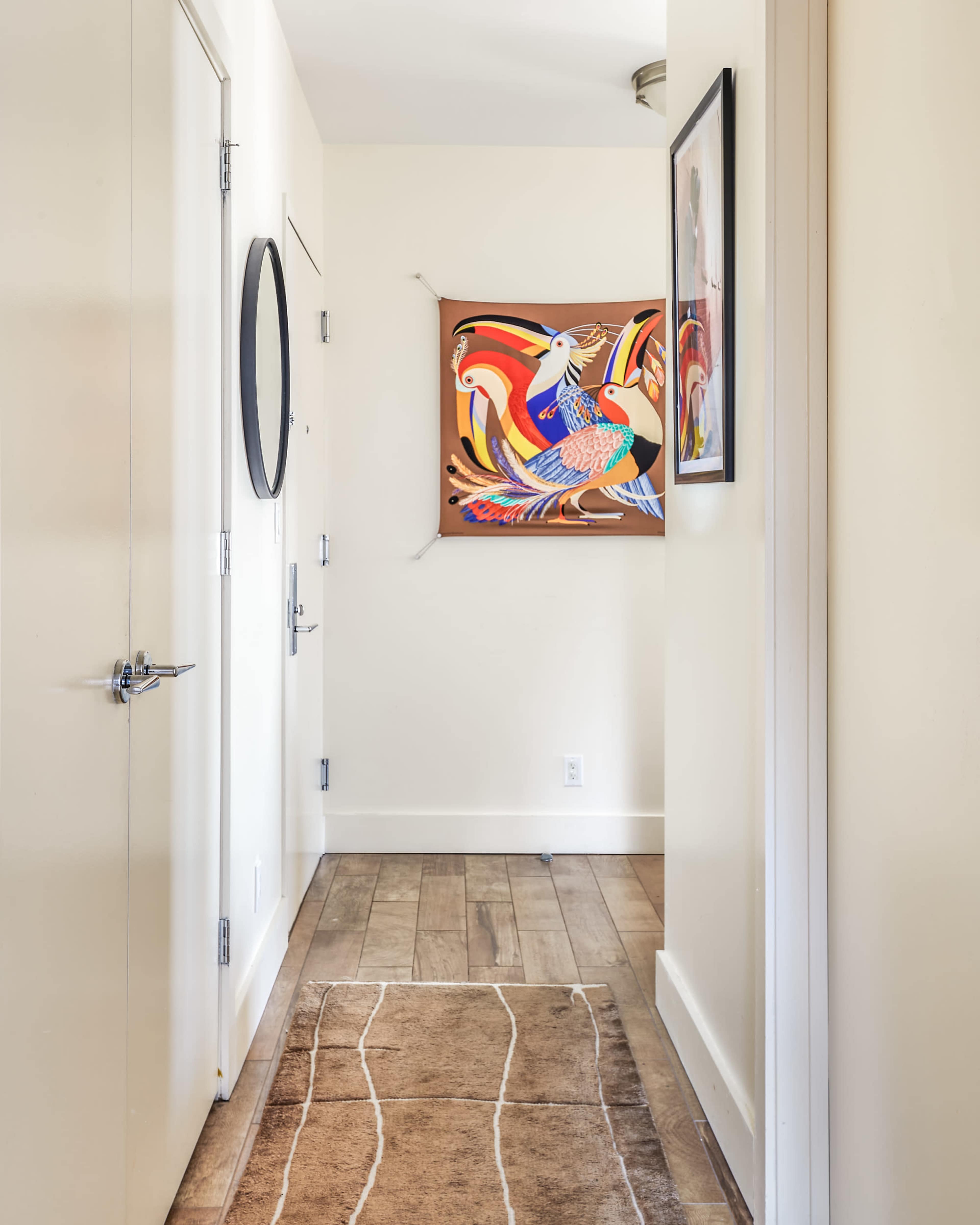 A hallway features a colorful artwork on the wall, a round mirror, and a wooden floor with a patterned rug.