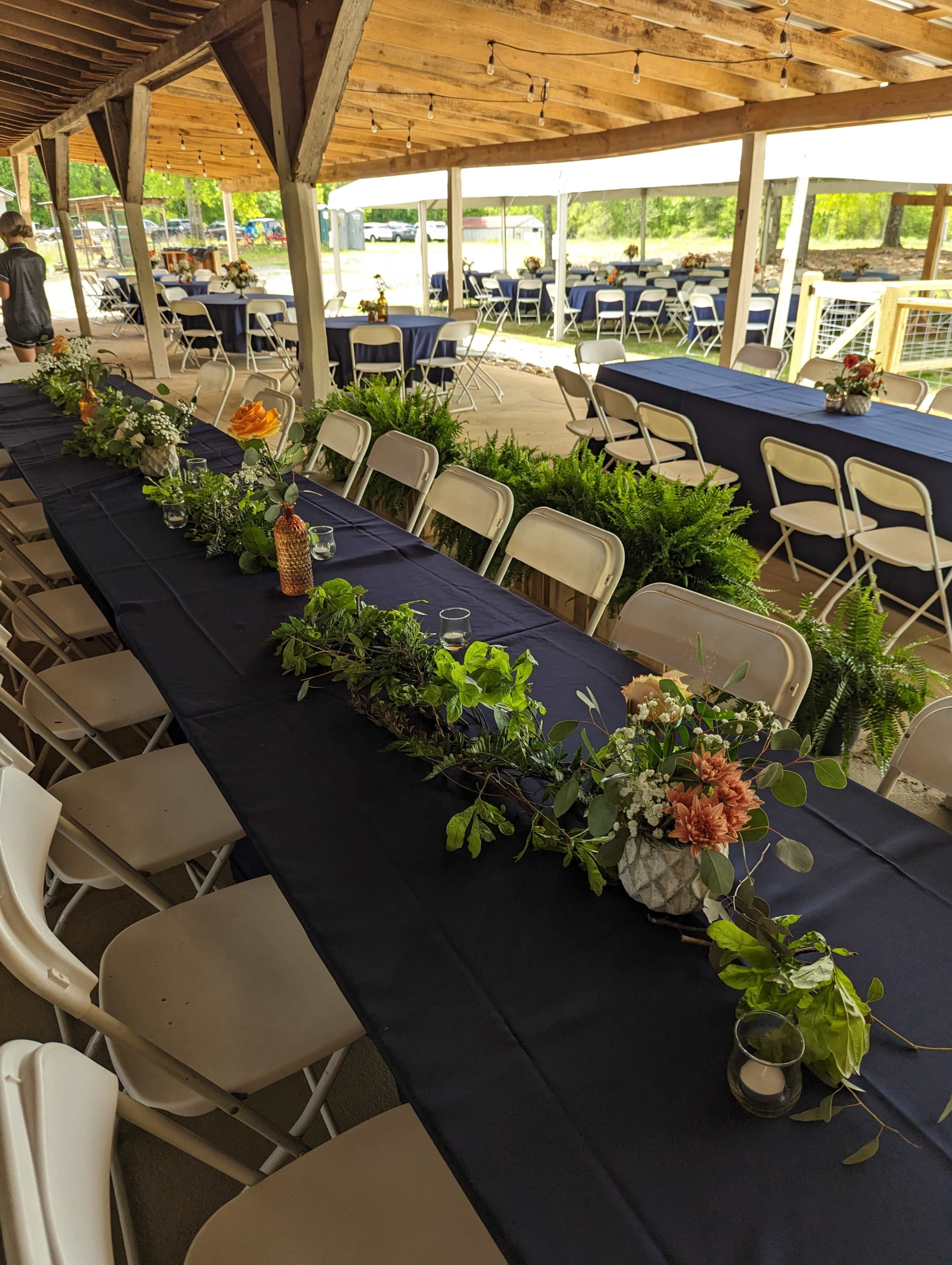 A decorated outdoor venue with long tables covered in dark cloth and adorned with greenery and floral centerpieces.