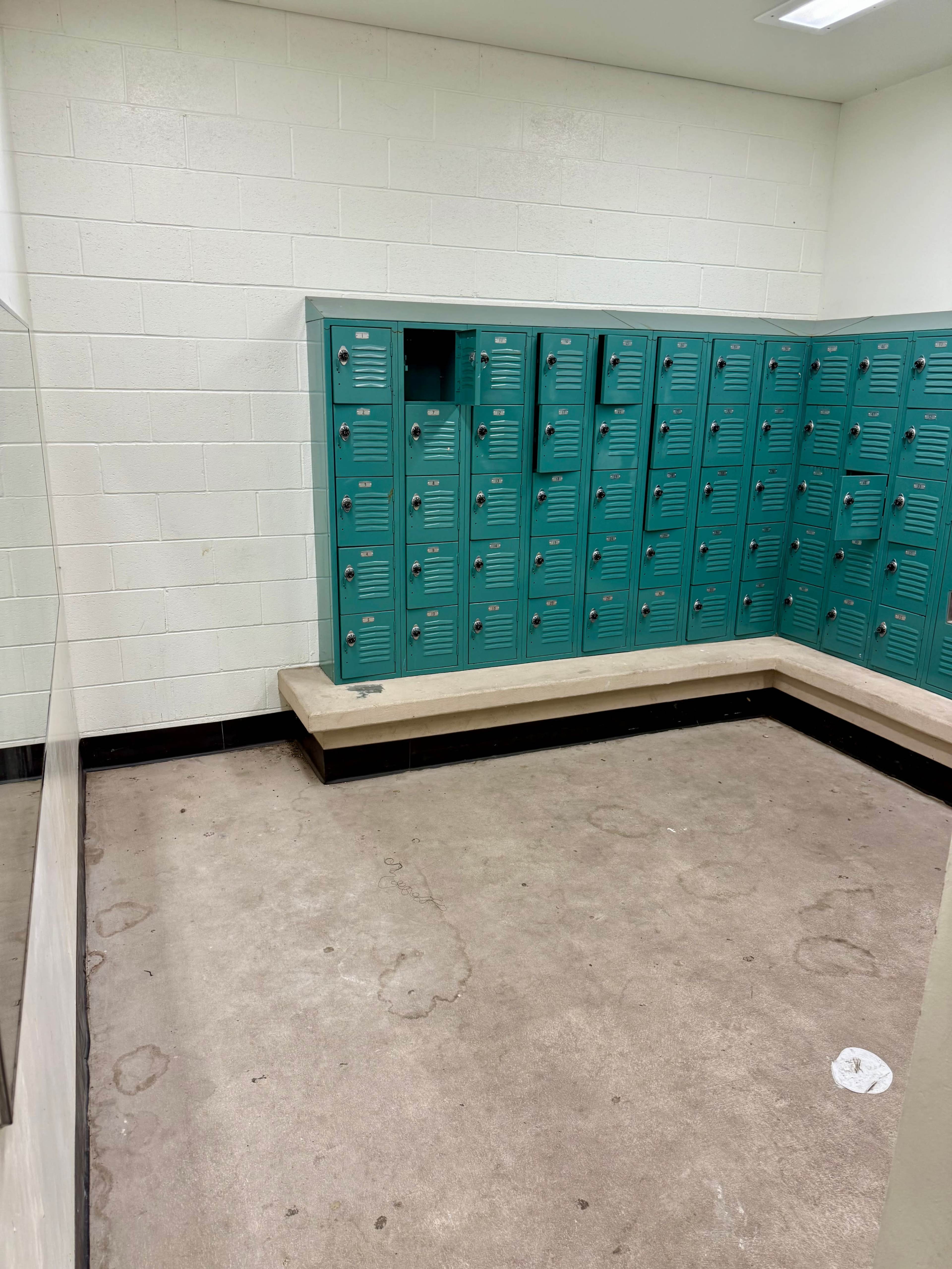 The image shows a vacant locker room with several teal lockers arranged against the wall and a bench along the side.