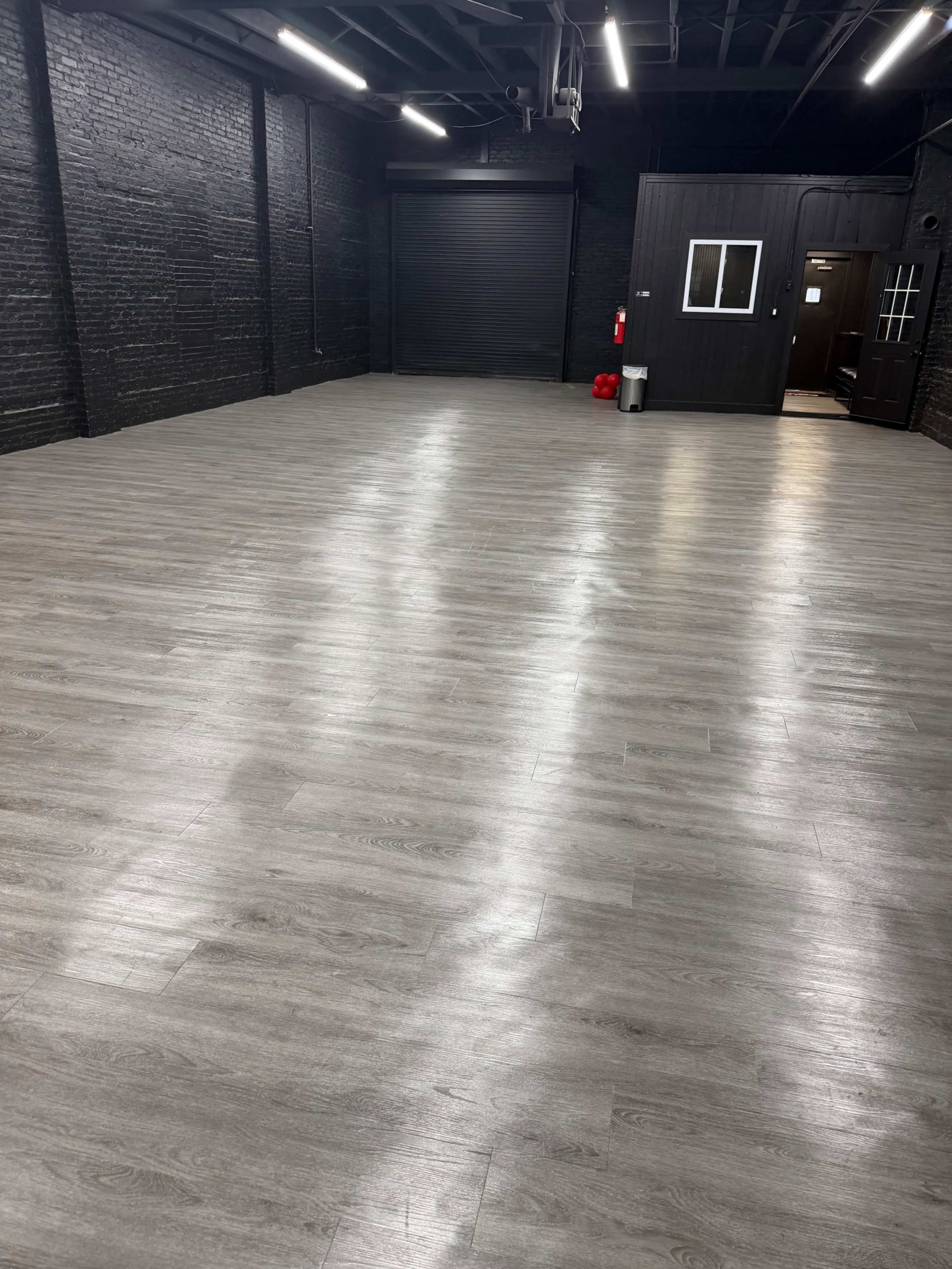 The image shows a spacious, empty room with a gray wood-like floor and black brick walls, featuring a closed door and a small window on one side.