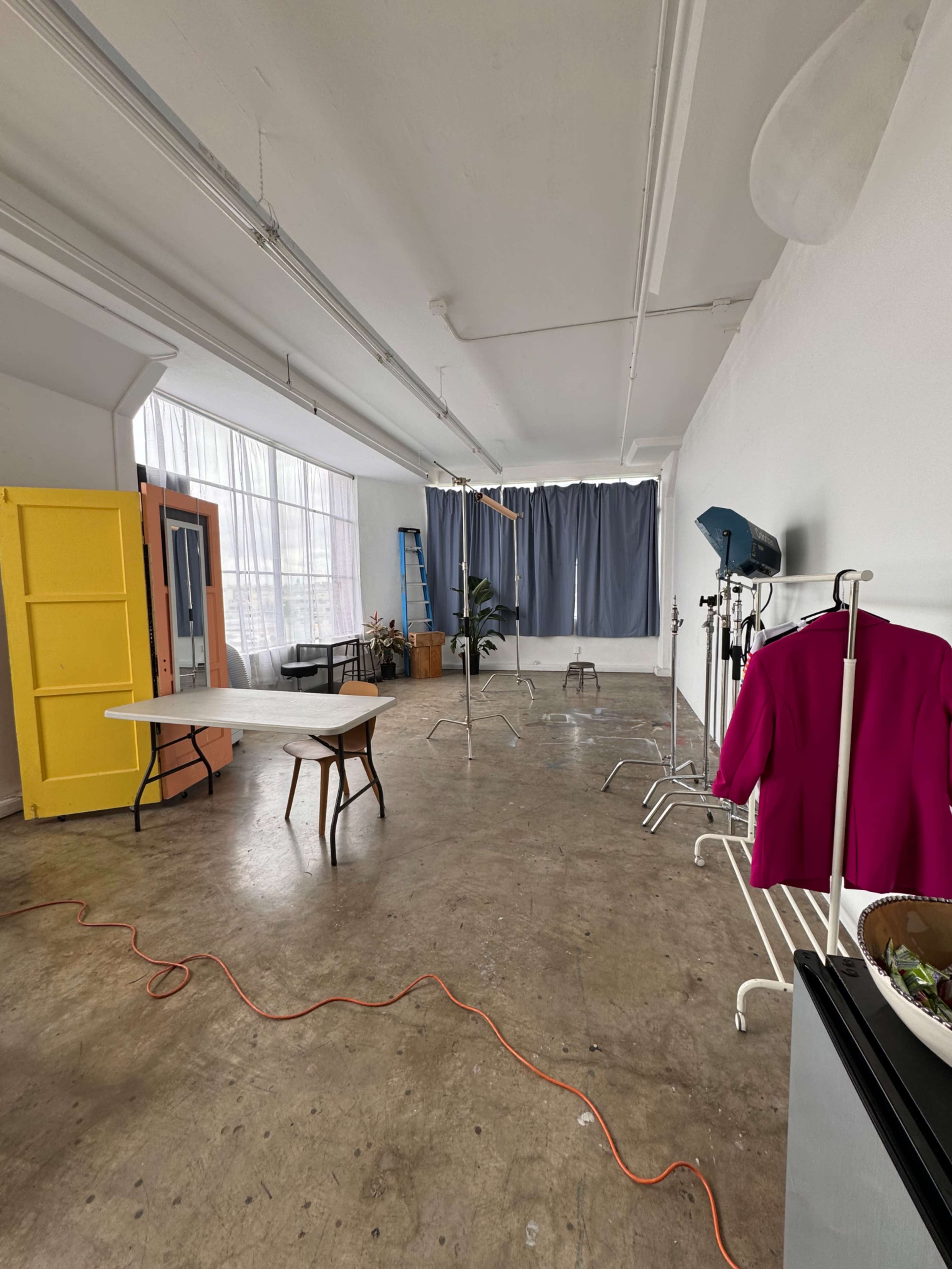 An empty studio space features a table, chairs, a yellow wardrobe, and clothes racks against a backdrop of gray curtains and large windows.