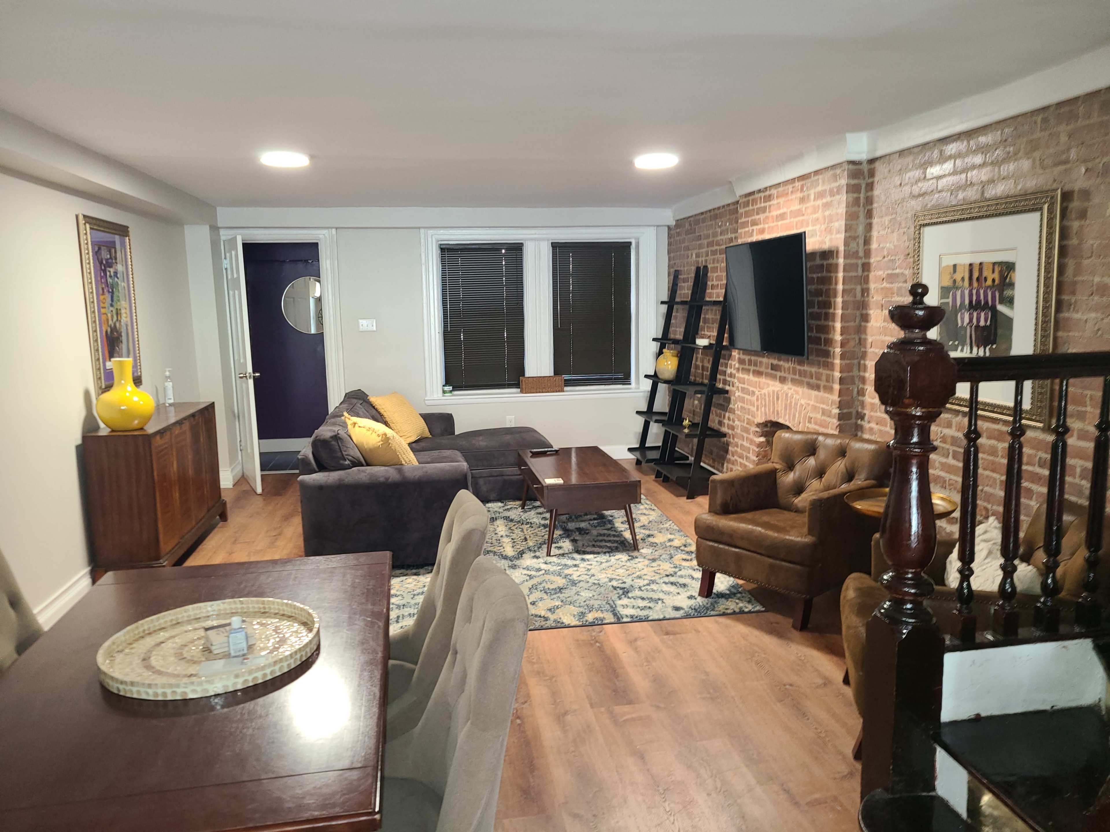 A modern living room with a brick wall, a sofa, armchair, coffee table, and dining area, illuminated by overhead lights.