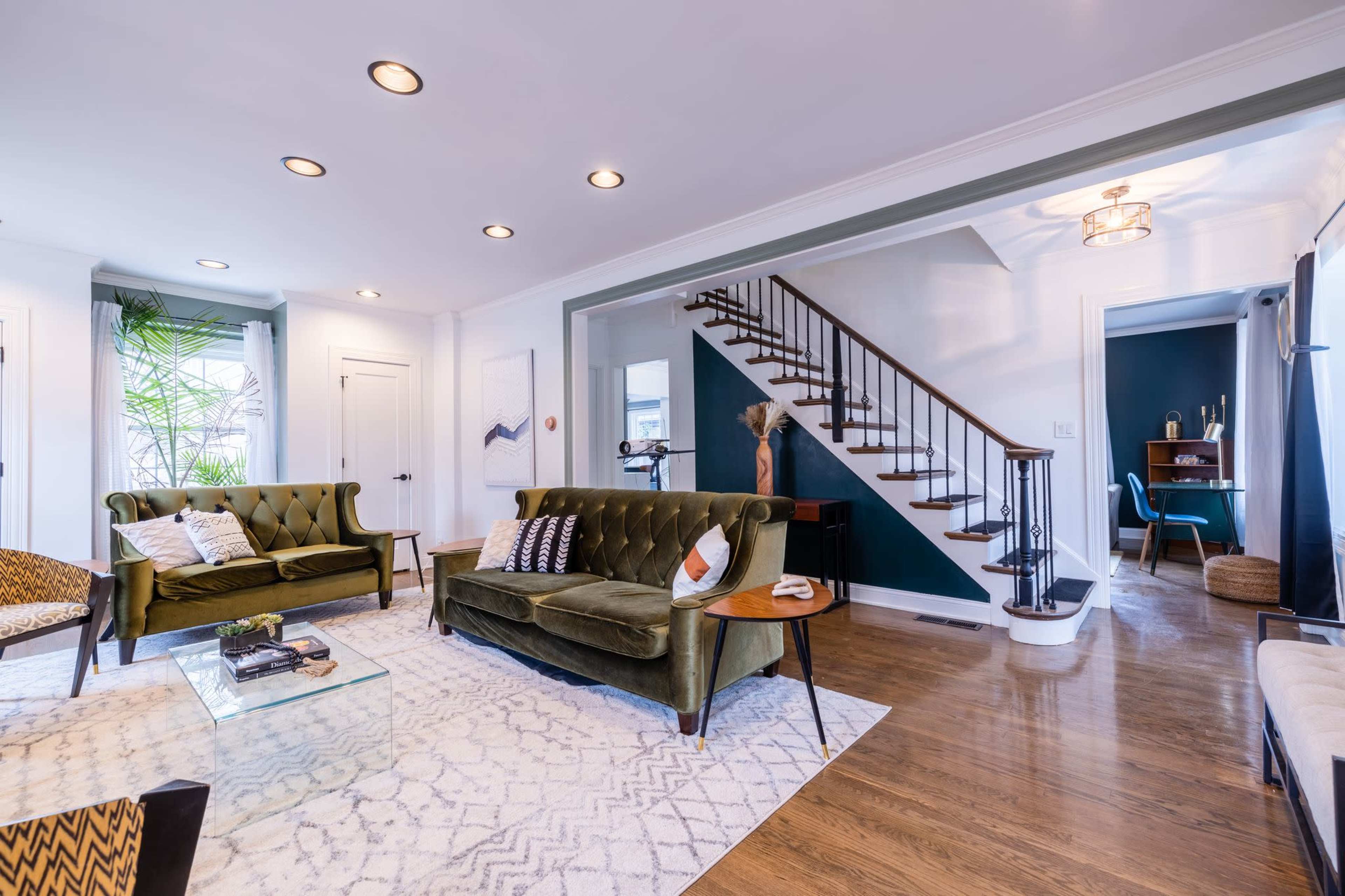 A stylish living room with green velvet sofas, a glass coffee table, and a staircase leading to an upper level.
