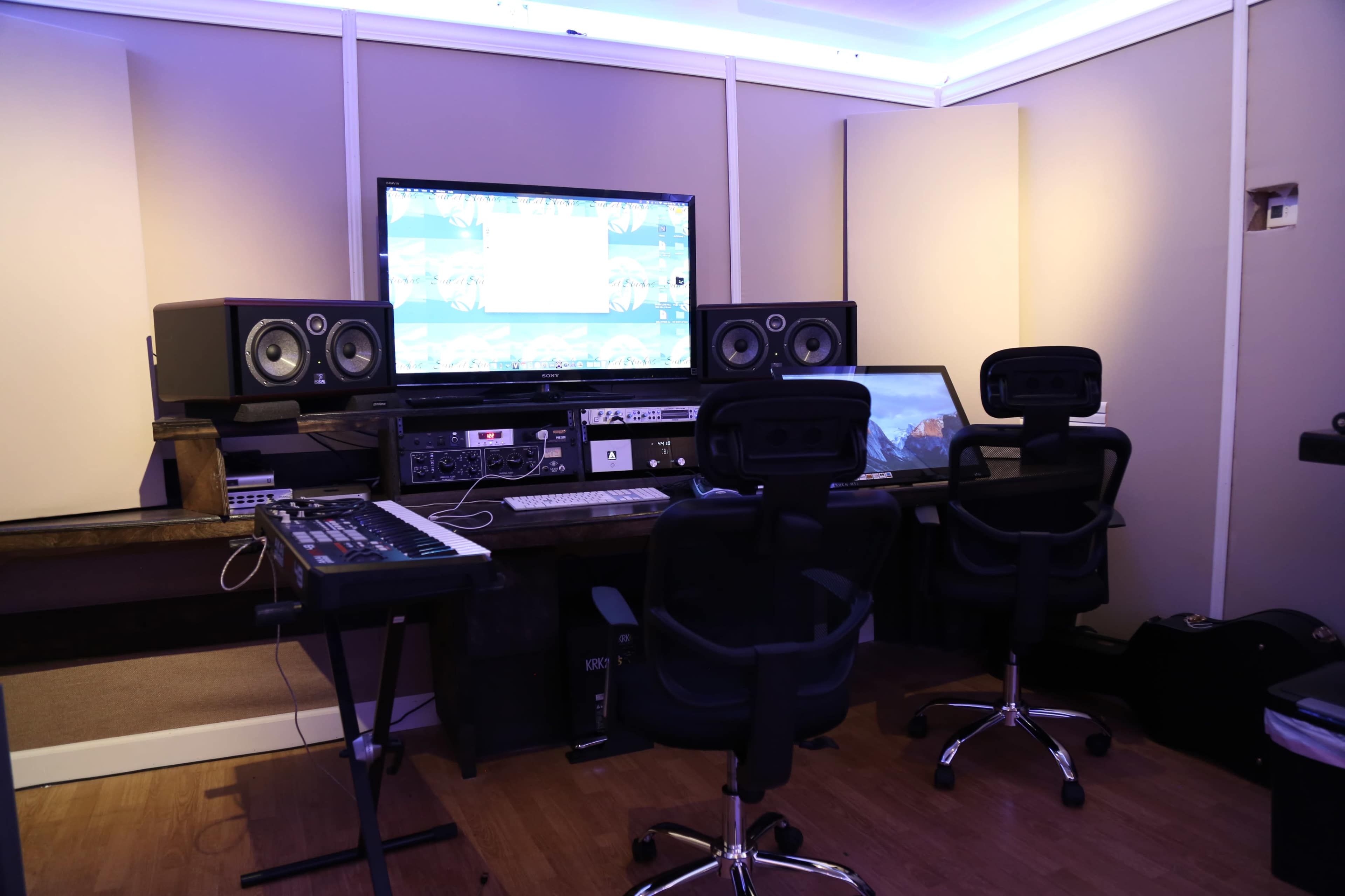 The image shows a modern music studio featuring dual computer monitors, audio equipment, and two ergonomic chairs.