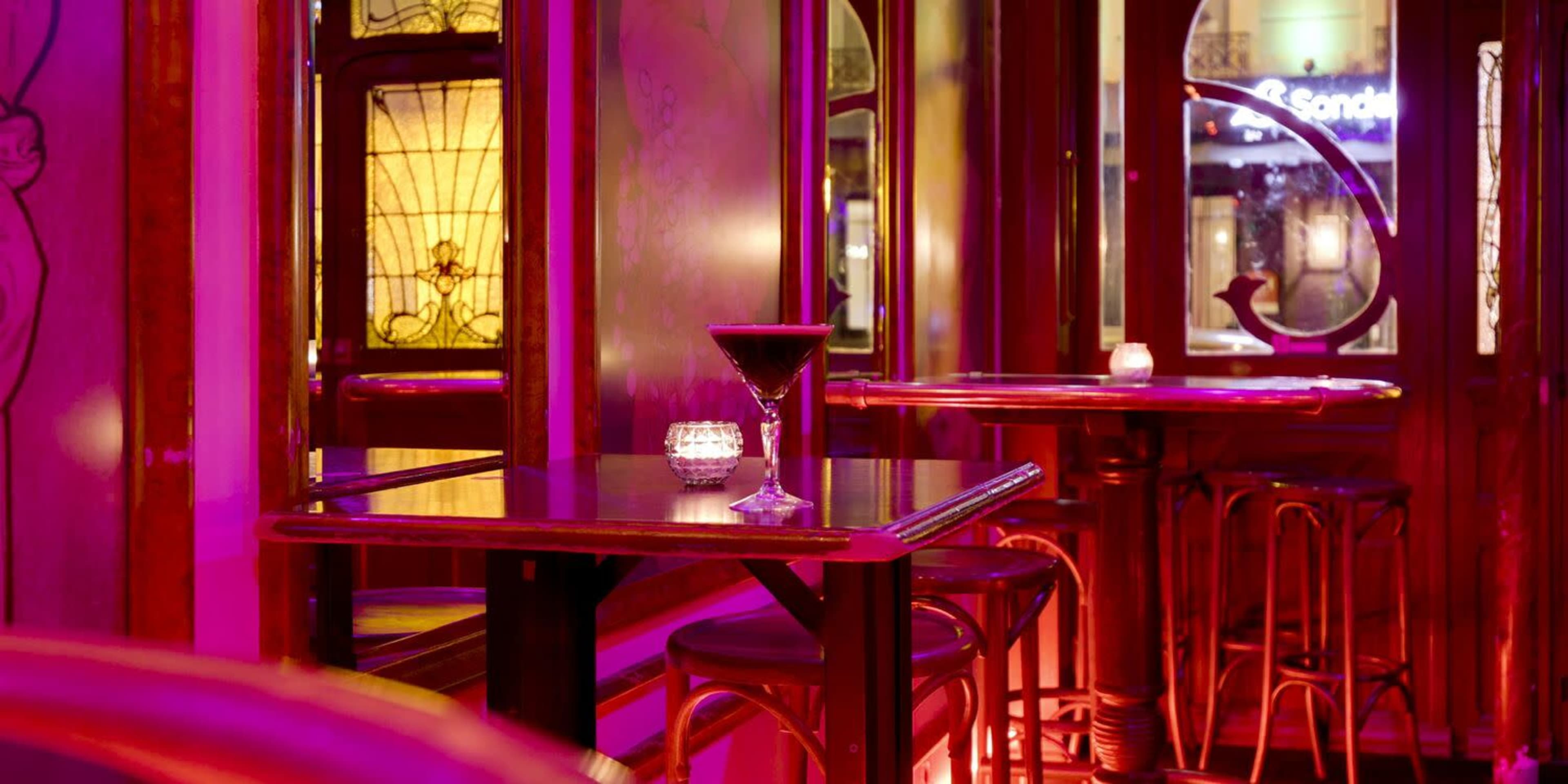 A dimly lit bar scene features a small table with a martini glass and a lit candle, surrounded by elegant wooden furniture and decorative glass elements.
