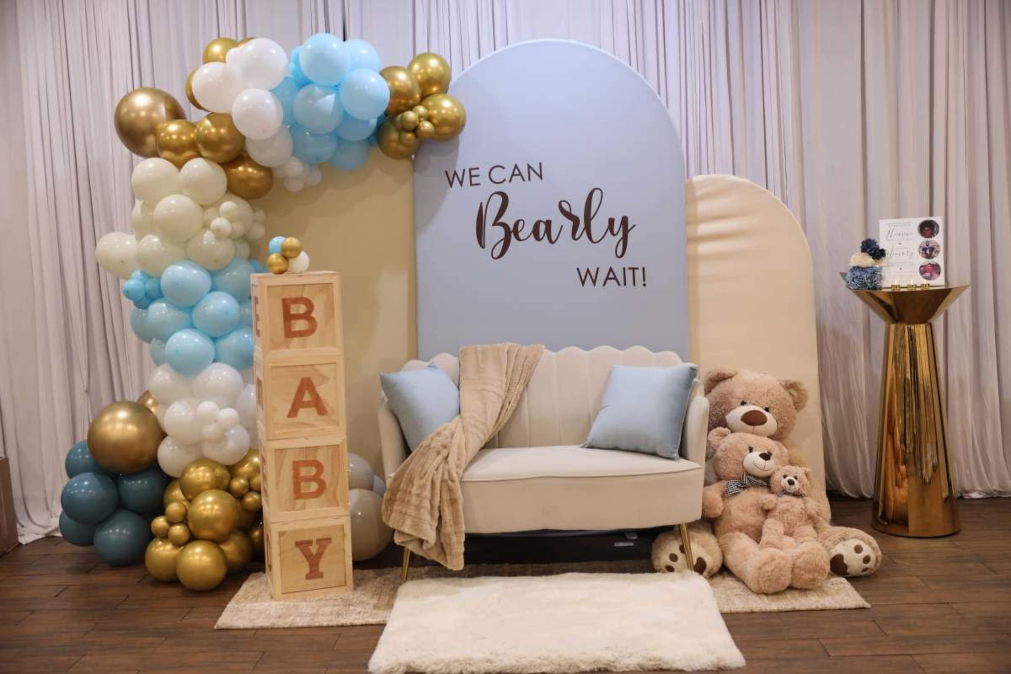 The image features a decorated baby shower area with a light blue backdrop, a plush white sofa, and balloon arrangements in blue, gold, and white, alongside large wooden blocks spelling "BABY" and plush teddy bears.