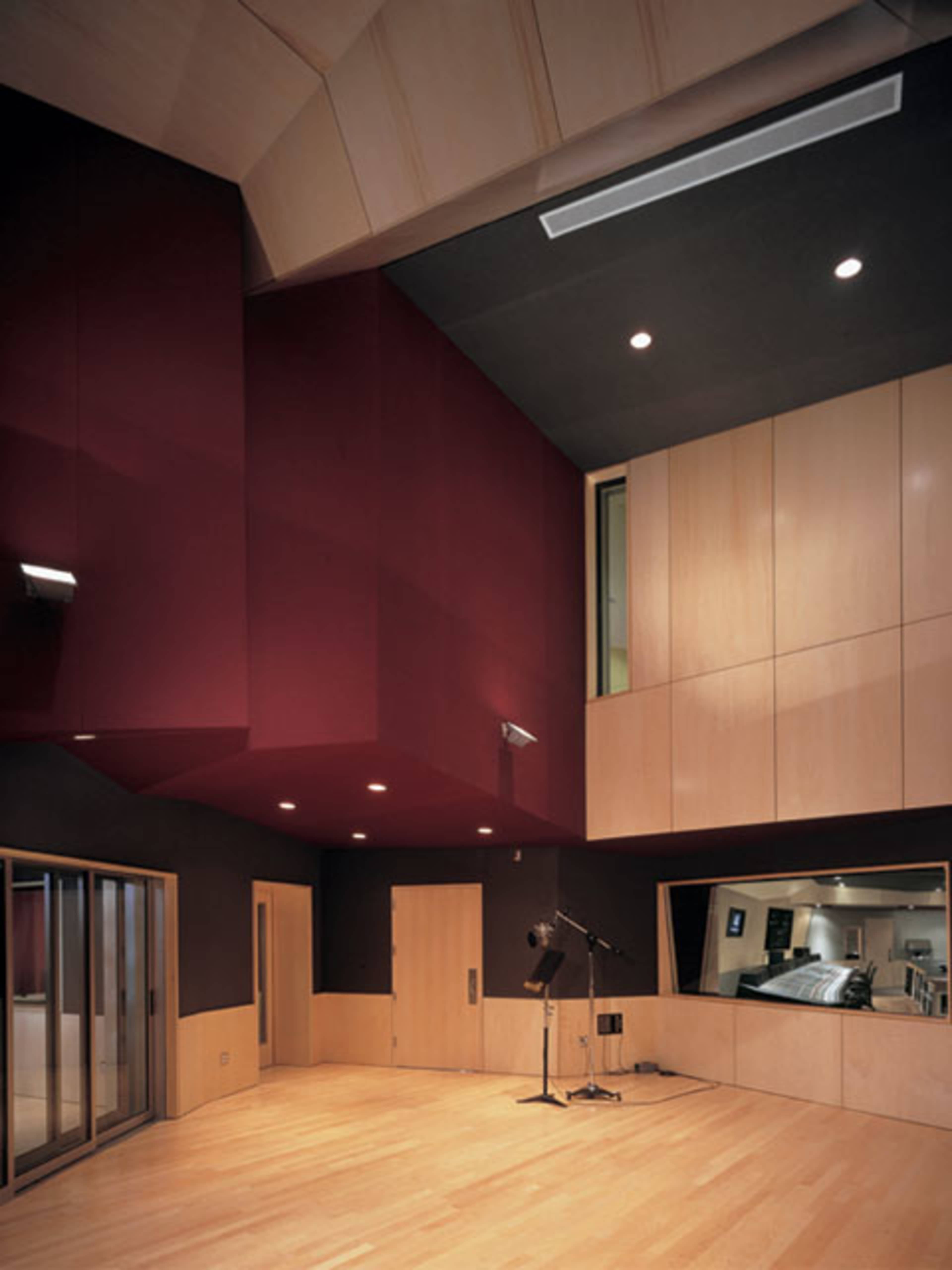 The image shows a modern recording studio with a wooden floor, red and black walls, and a large window overlooking another room.