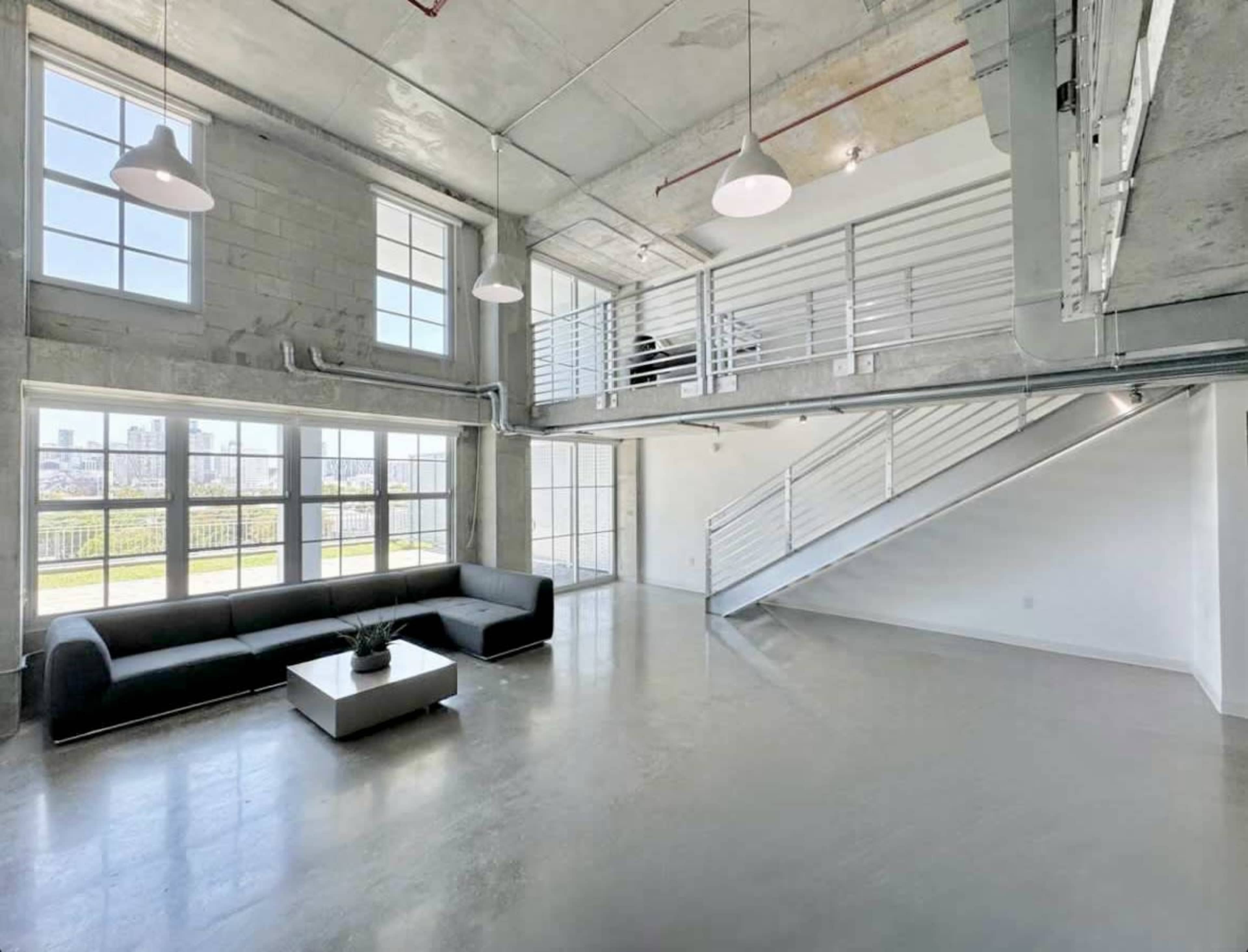A modern industrial-style open space with large windows, a sofa, a coffee table, and a staircase leading to a loft area.