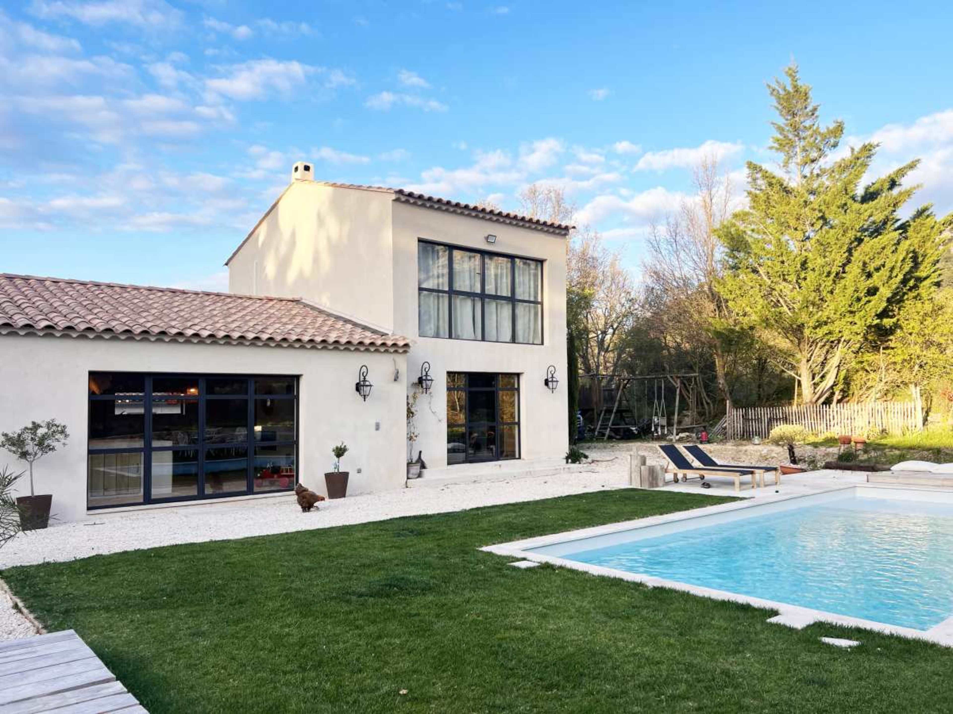 A modern two-story house with large windows is surrounded by a neatly landscaped yard featuring a swimming pool and a lounge chair.