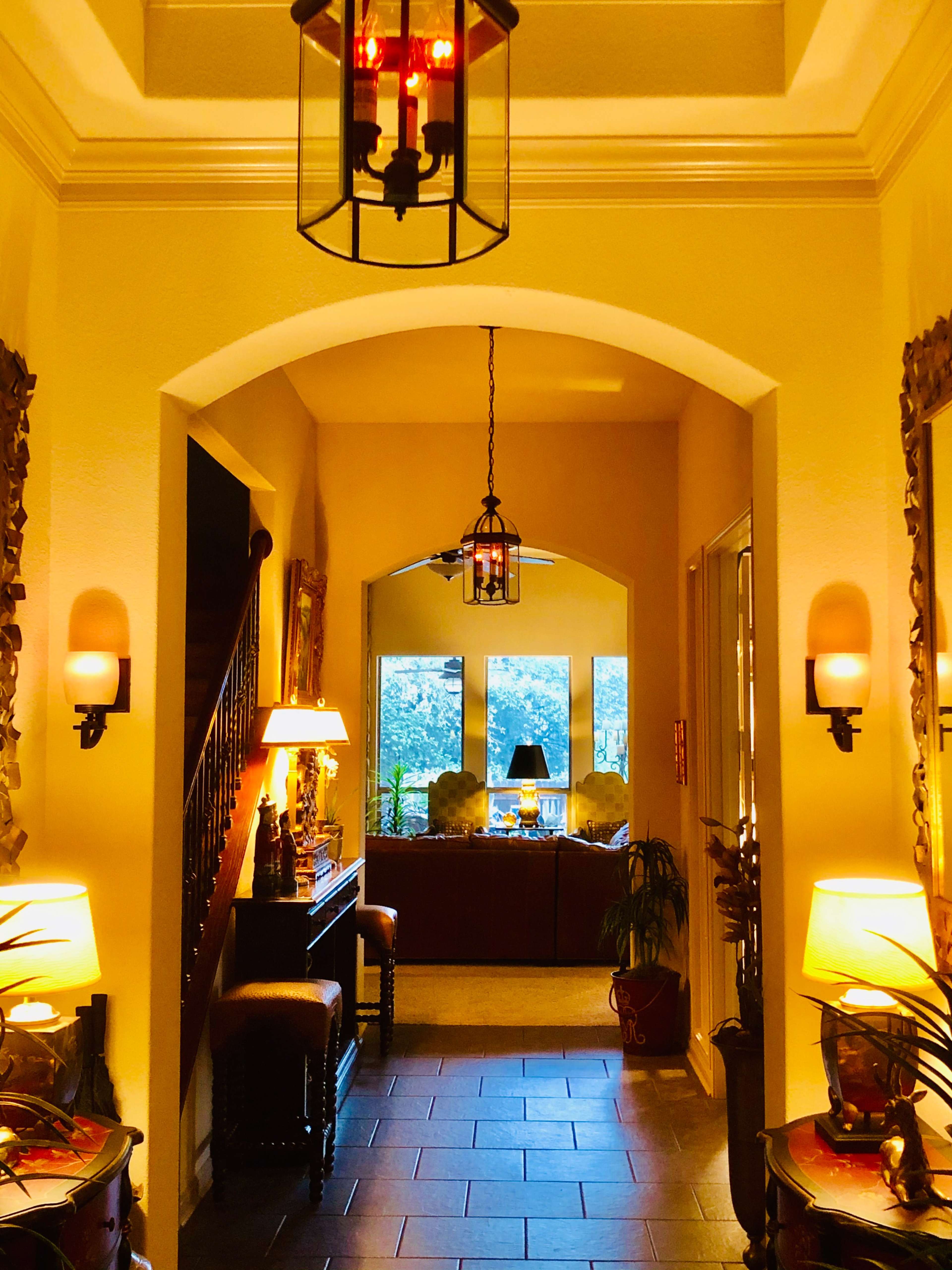 A well-lit hallway with arched doorways, featuring decorative lamps, potted plants, and a view into a furnished room at the end.