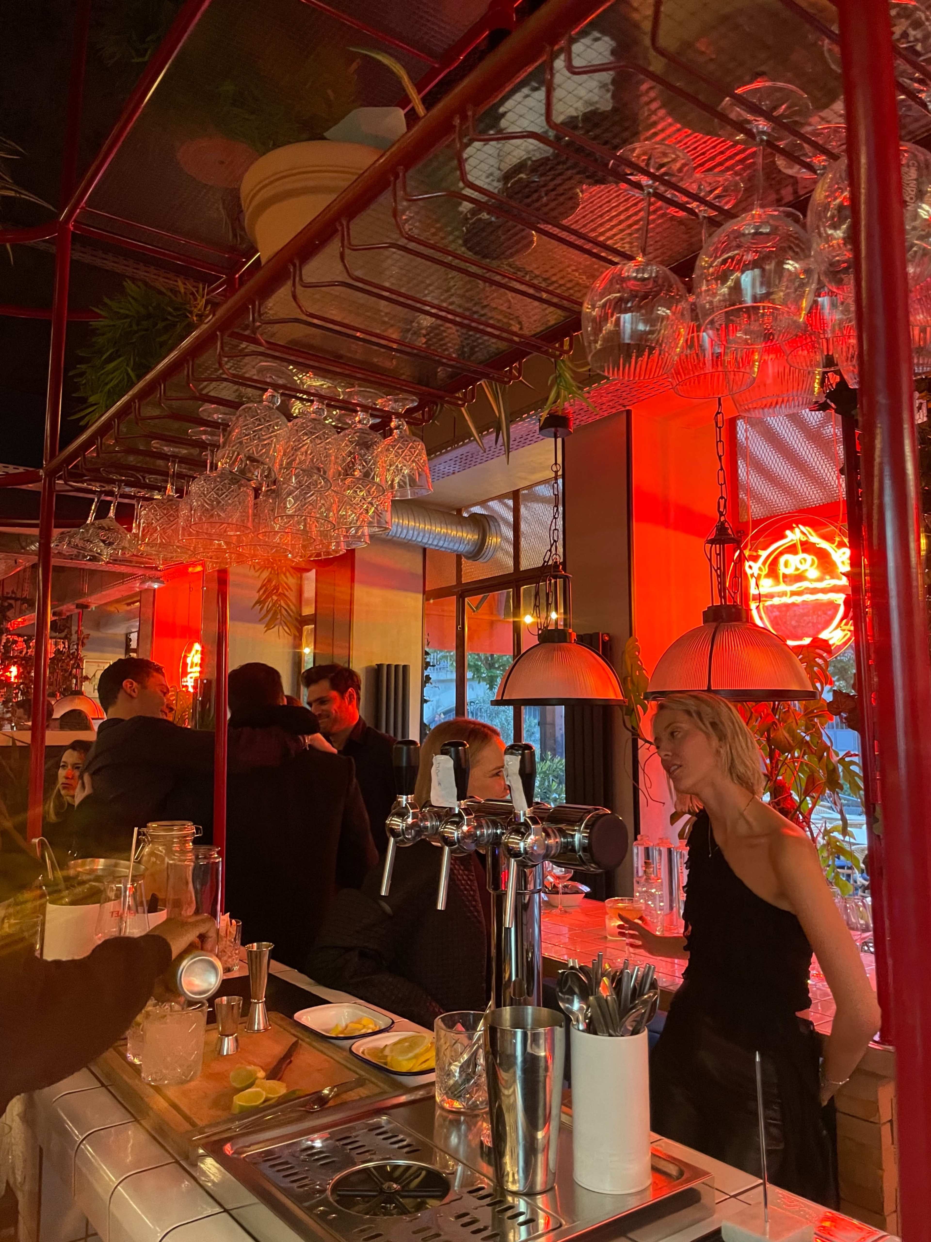 A dimly lit bar scene with patrons gathered around the counter, illuminated by red neon lights and glassware hanging above.