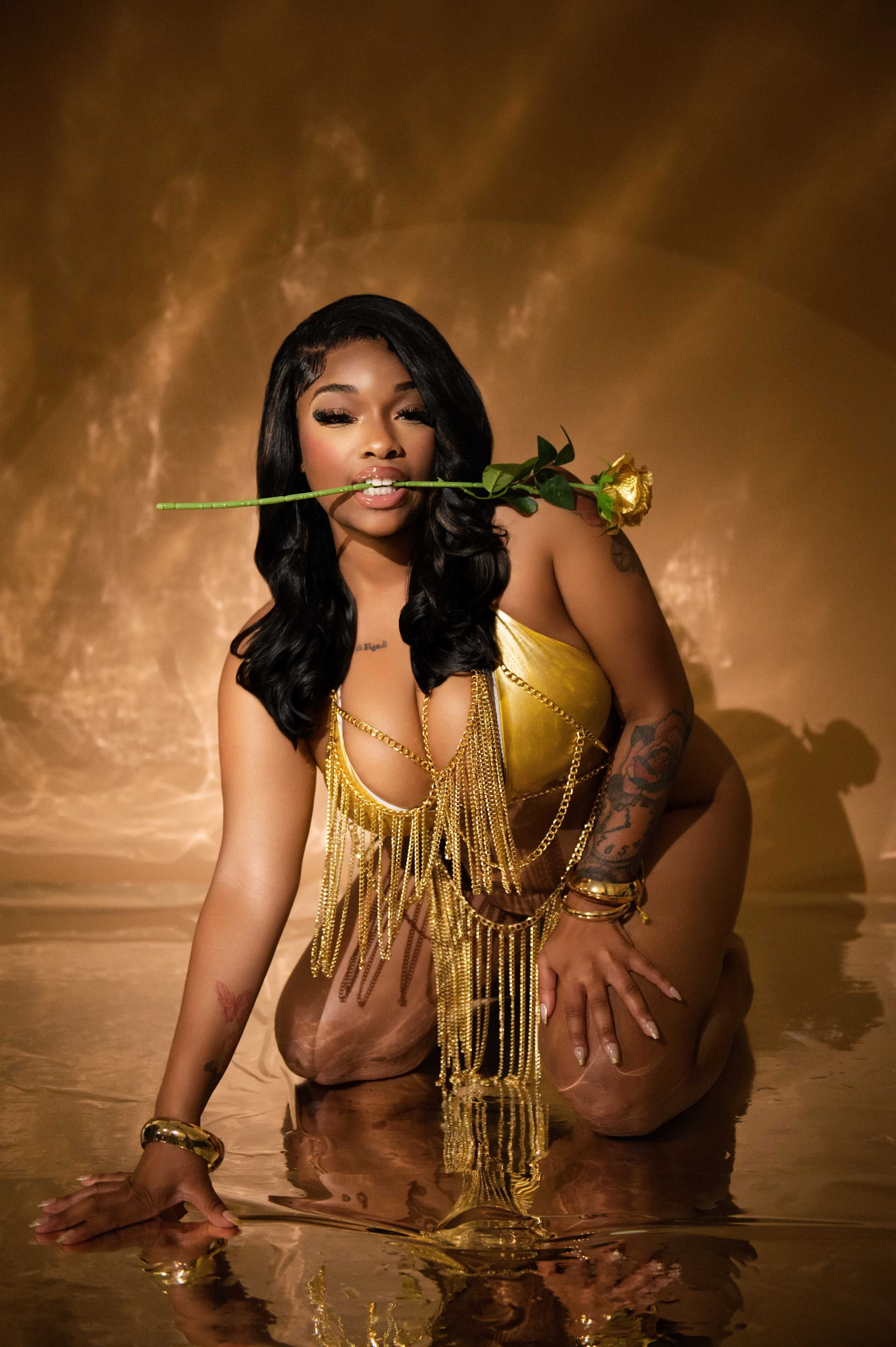 A woman in a gold bikini and jewelry poses in water with a yellow rose in her mouth, set against a golden background.