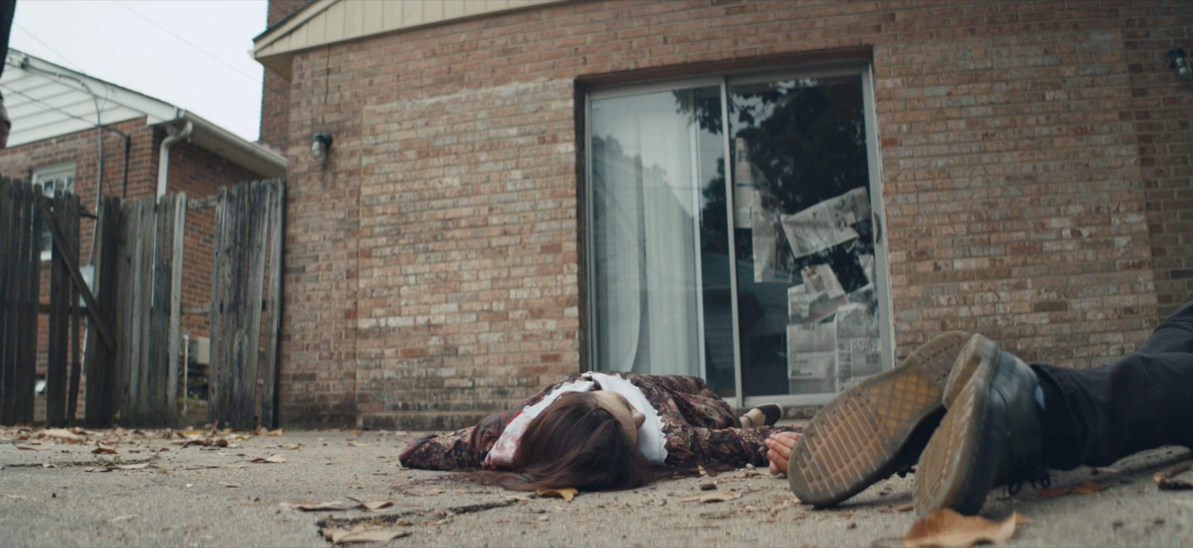 A person lies on the ground beside a house, appearing unresponsive, with scattered leaves around them.