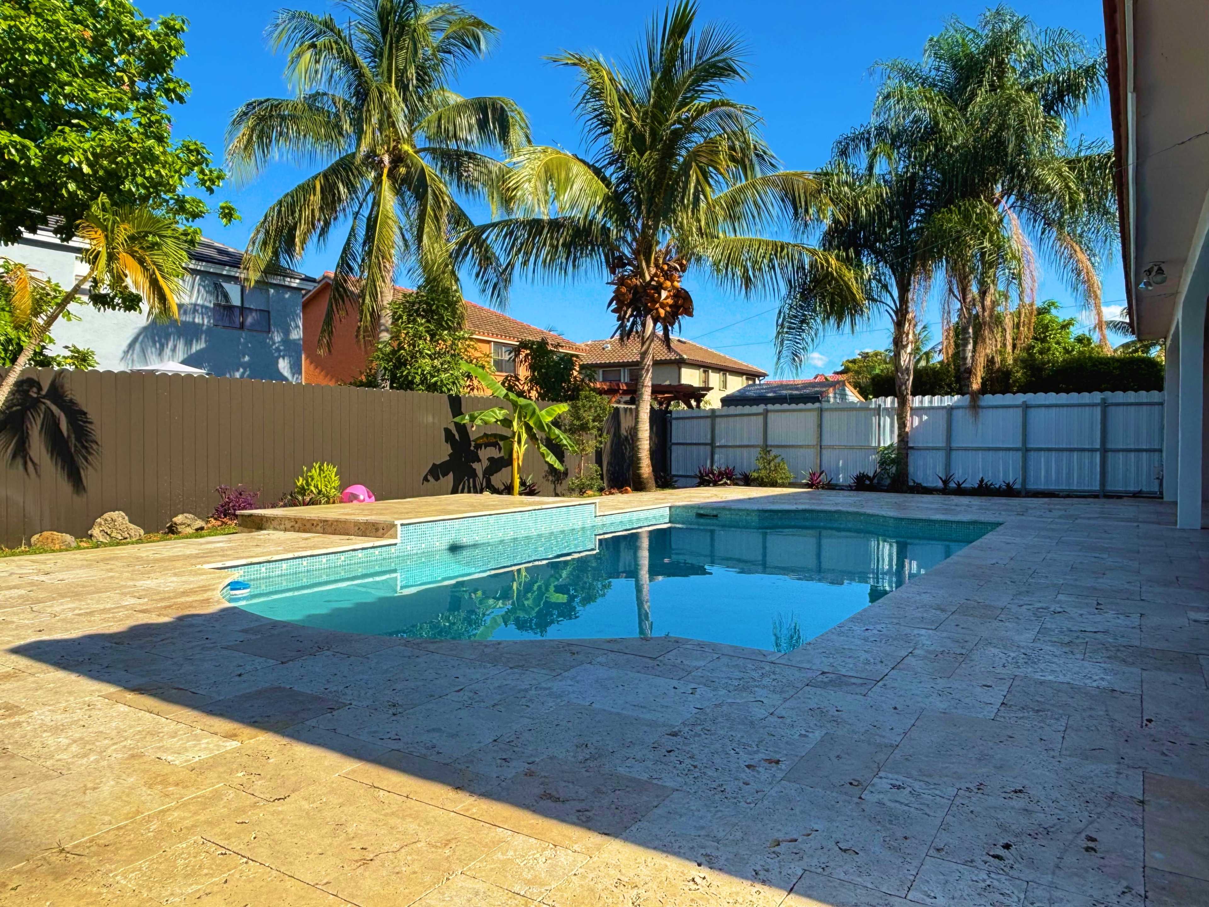 The image depicts a backyard pool area surrounded by palm trees and a garden, with a clear blue sky above.