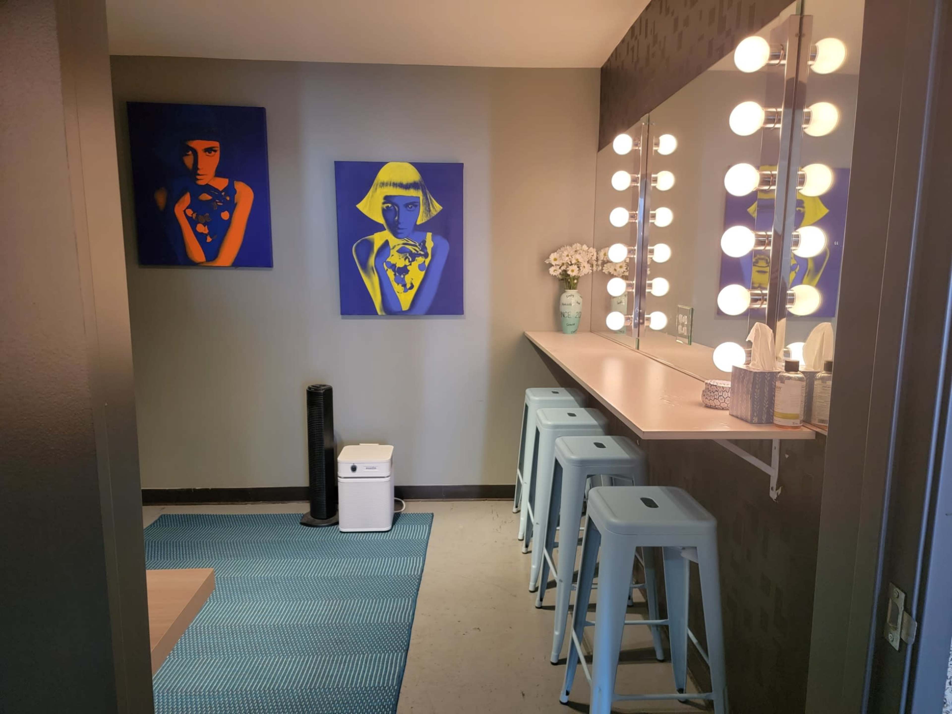 A bathroom with two blue art pieces on the wall, a vanity area with round light bulbs, a small table with stools, and a rug on the floor.