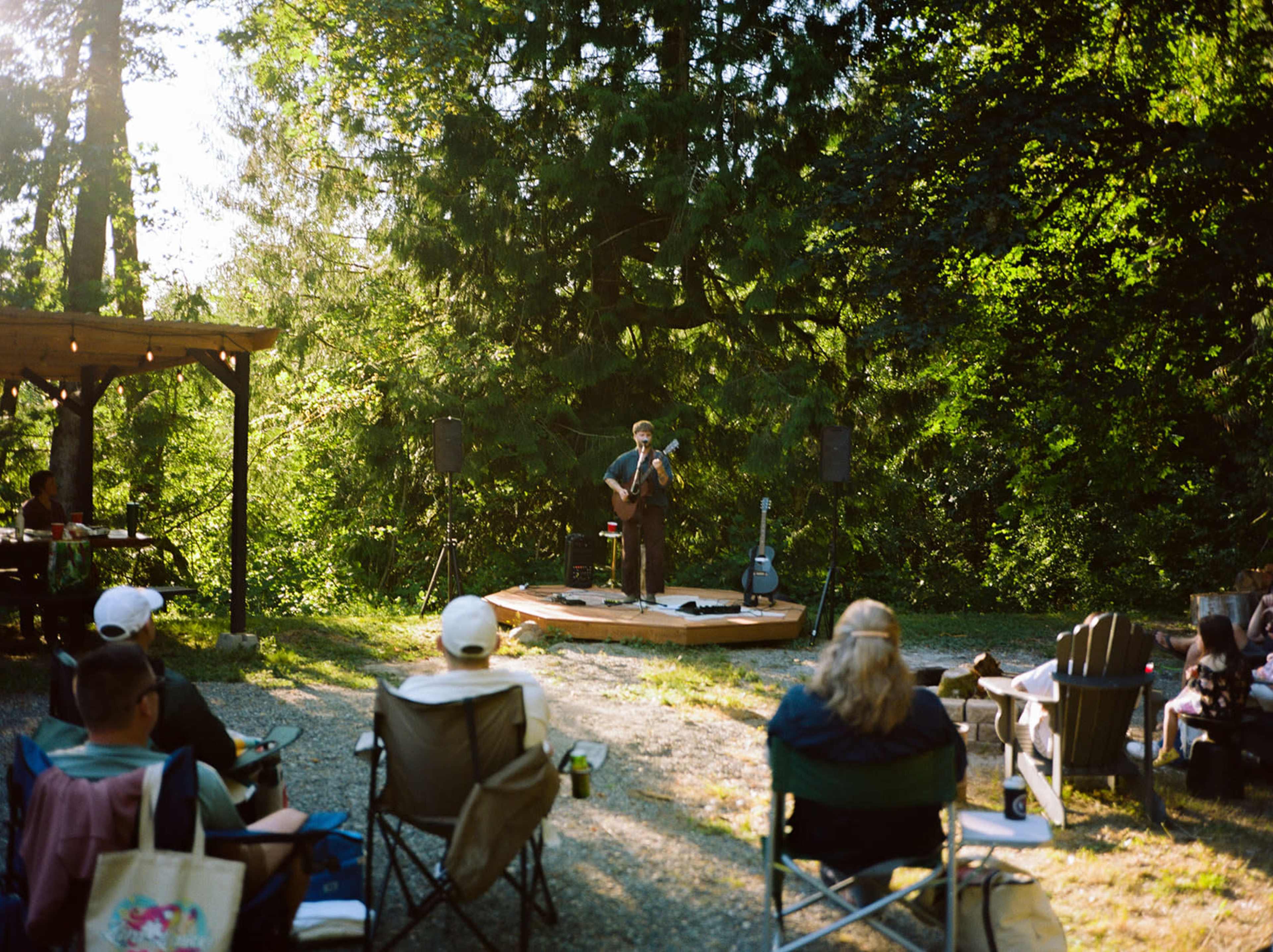 A musician performs on a wooden stage surrounded by an audience seated in lawn chairs under tall trees.