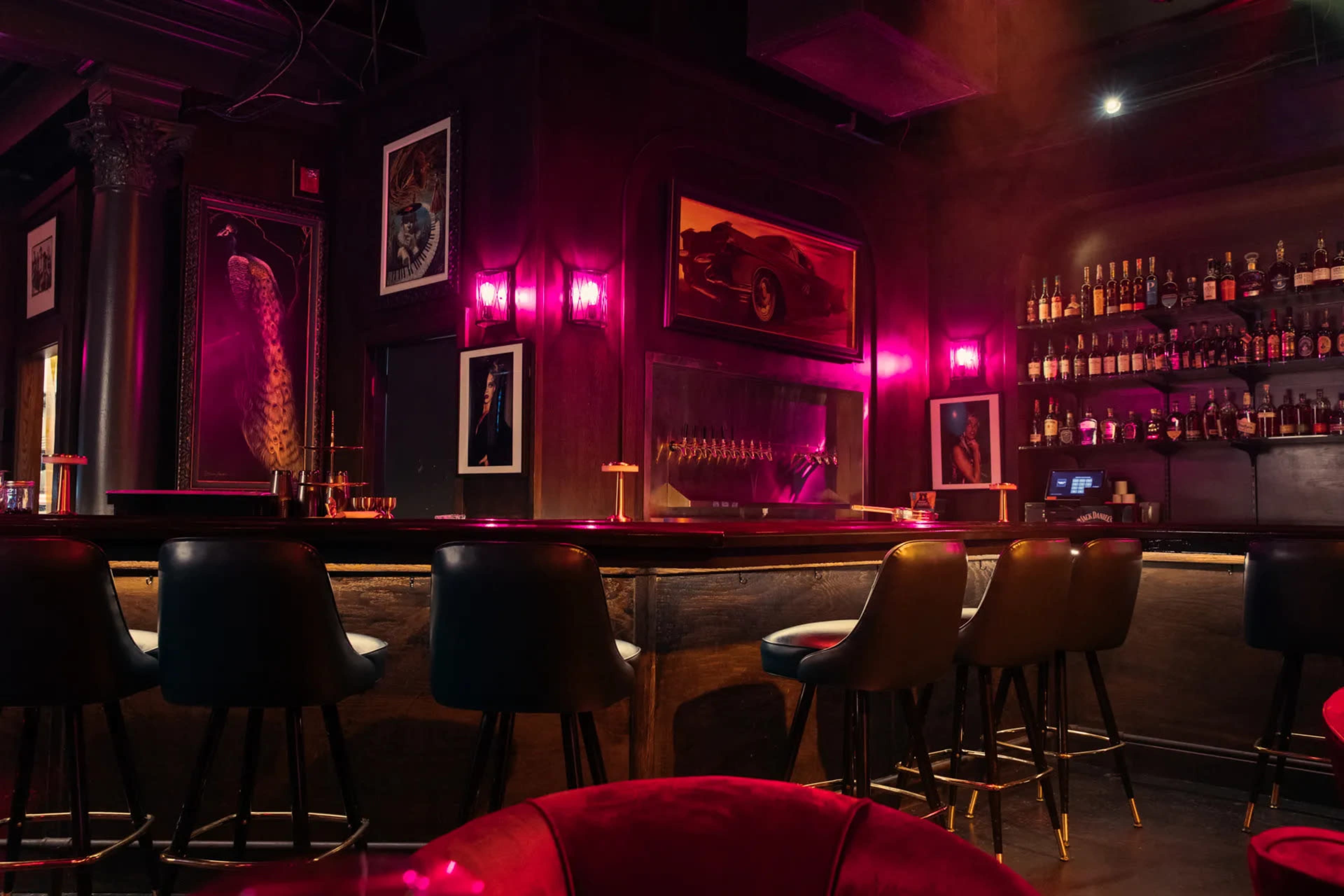 The image shows a dimly lit bar with a long counter, high-backed chairs, and shelves lined with bottles.
