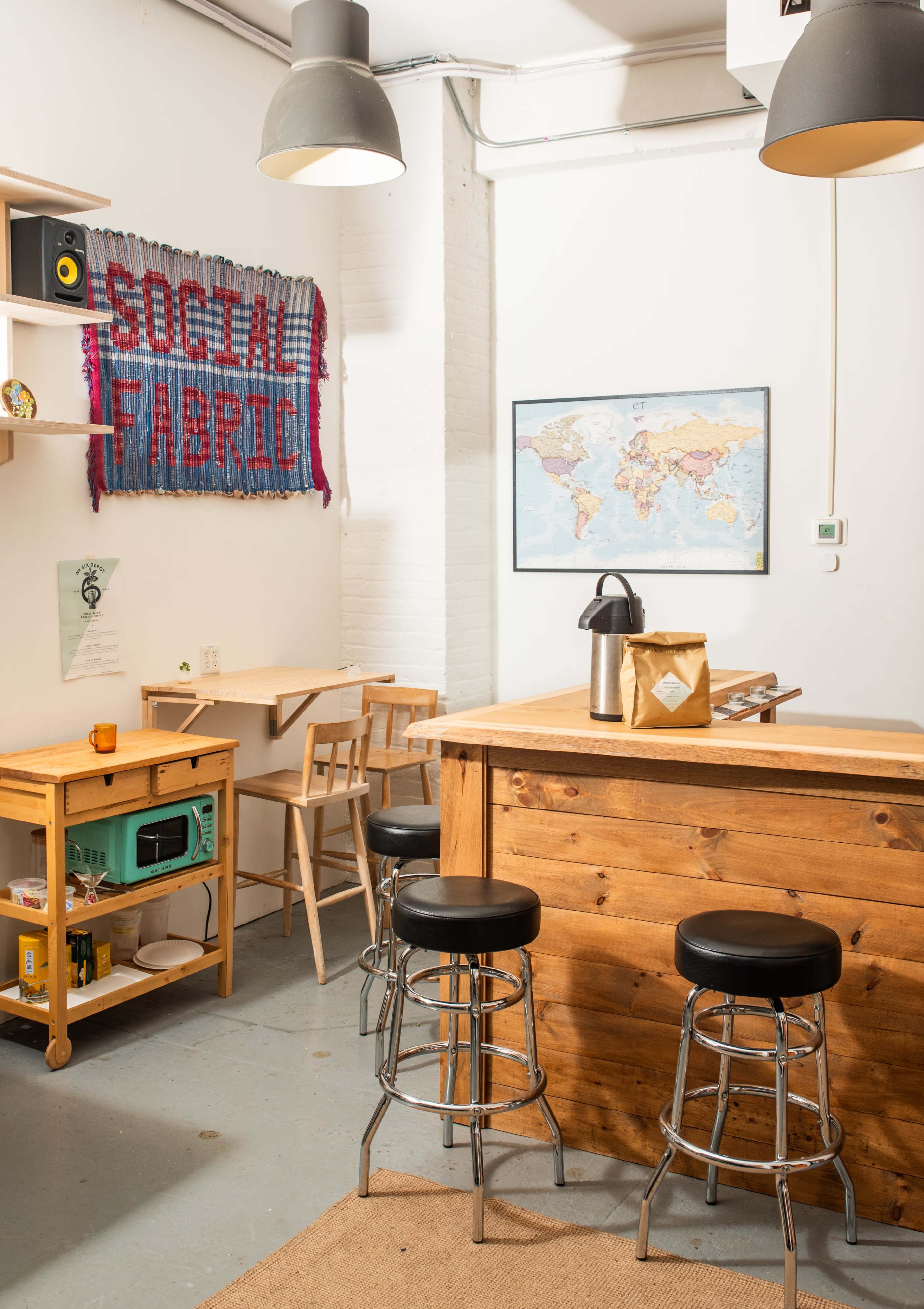 The image shows a cozy workspace with wooden furniture, bar stools, a world map on the wall, and decorative items including a woven tapestry that reads "SOCIAL FABRIC."