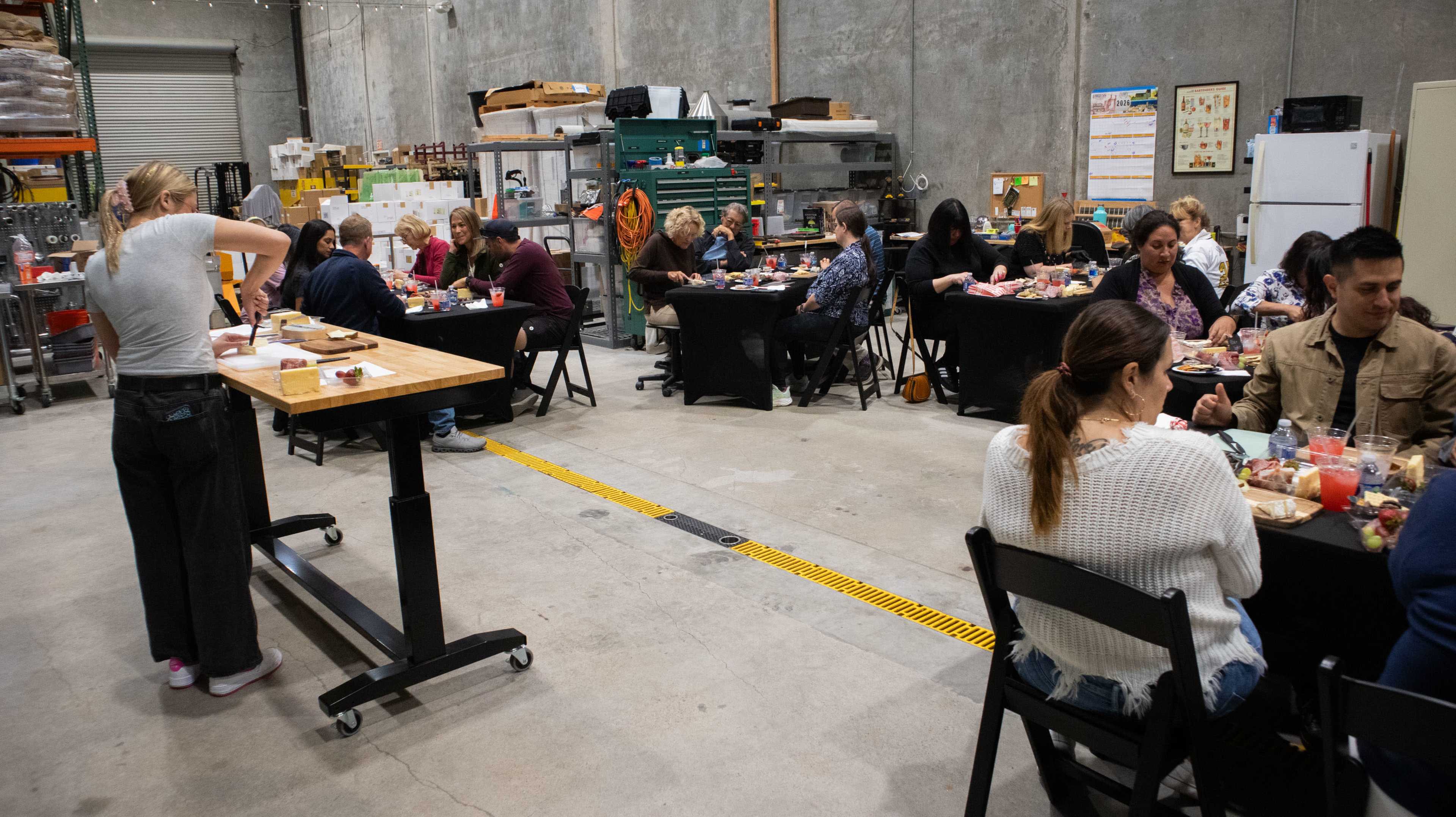 A group of people dine on meals at tables in a warehouse setting, while another person prepares food at a counter.