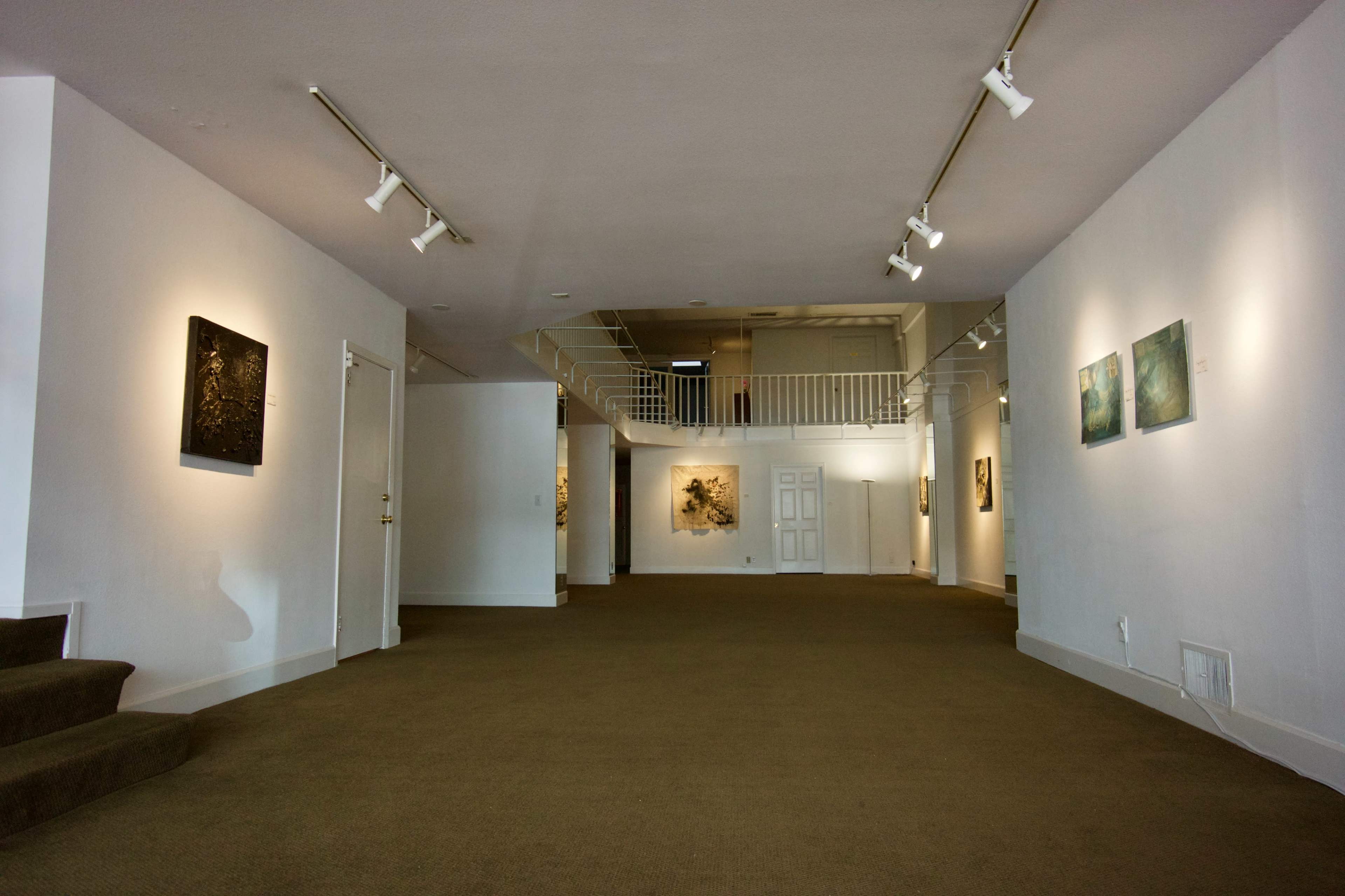 The image shows the interior of an art gallery, featuring various artworks displayed on the walls and a staircase leading to an upper level.