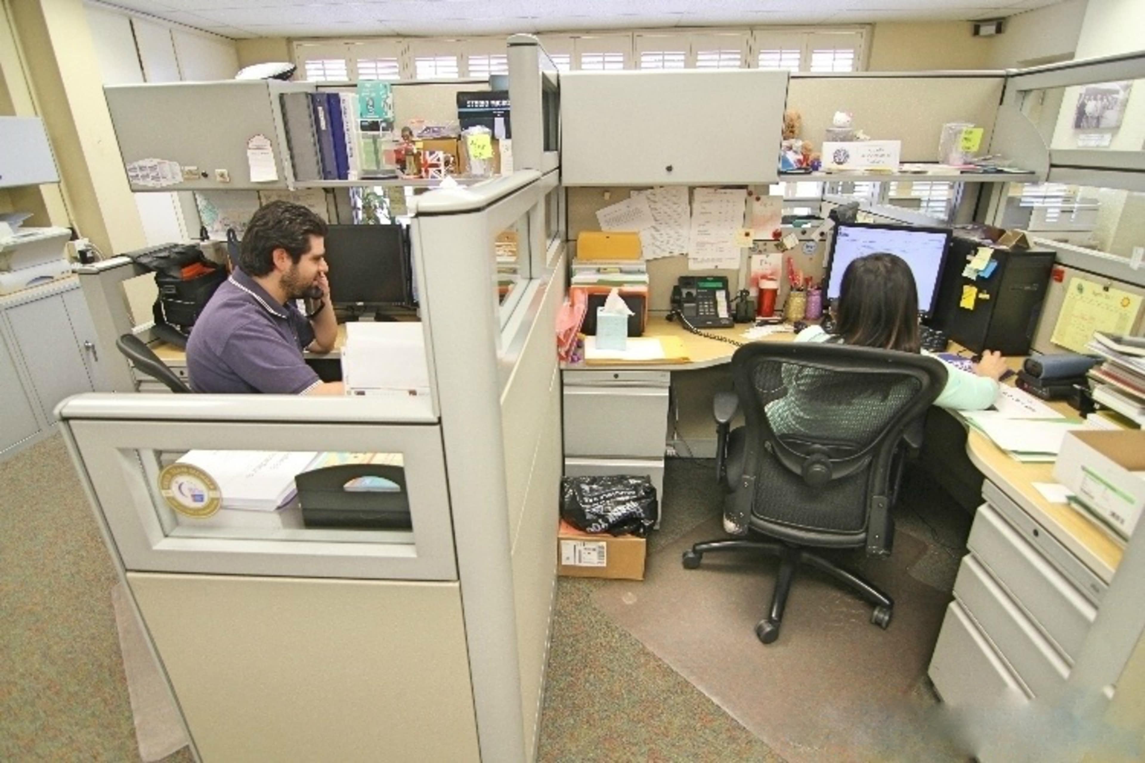 Two office workers are seated at adjacent cubicles, one talking on a phone and the other focused on a computer screen.