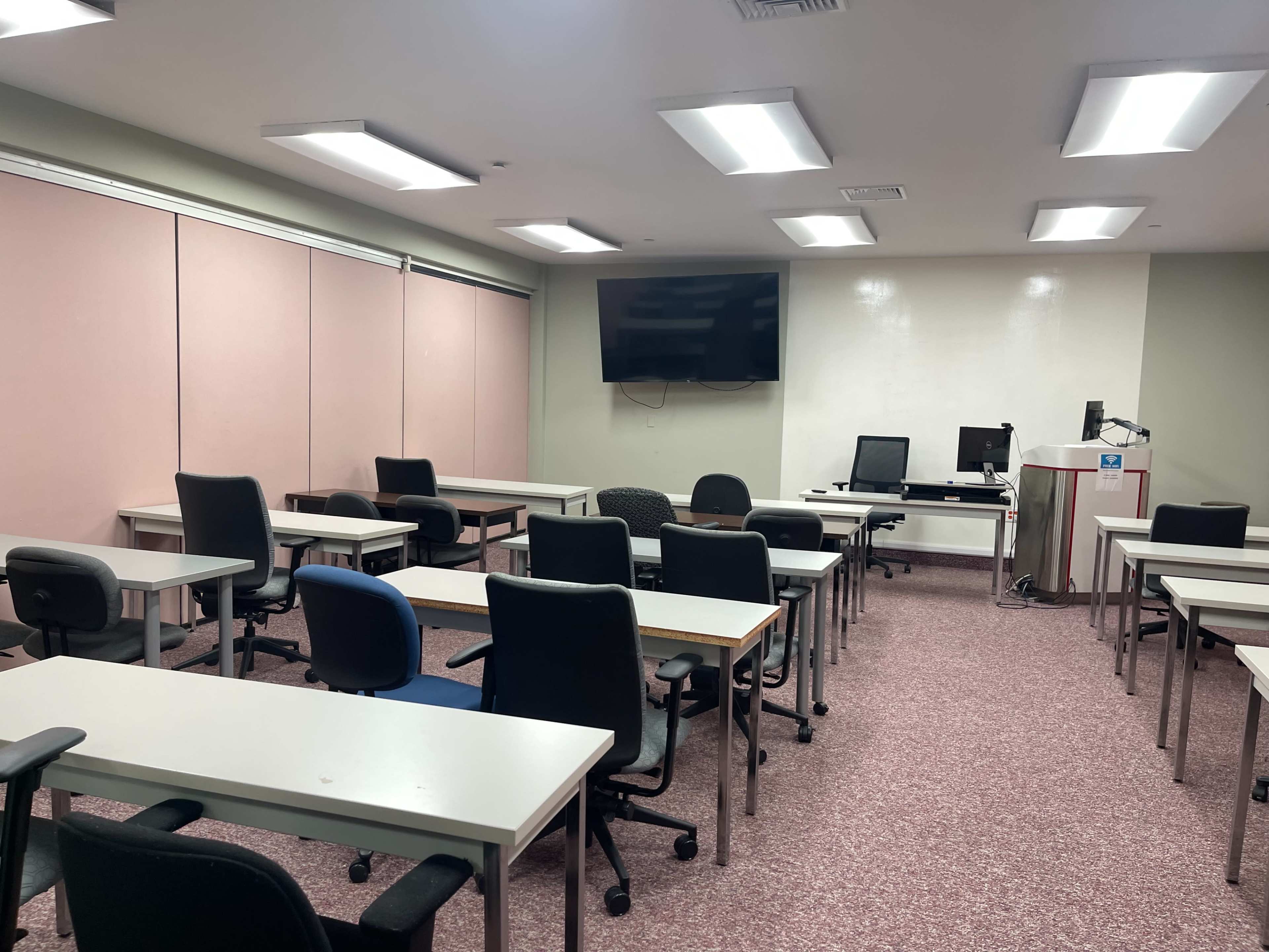 The image shows a classroom with rows of desks and chairs, a large monitor on the wall, and a teacher's station at the front.