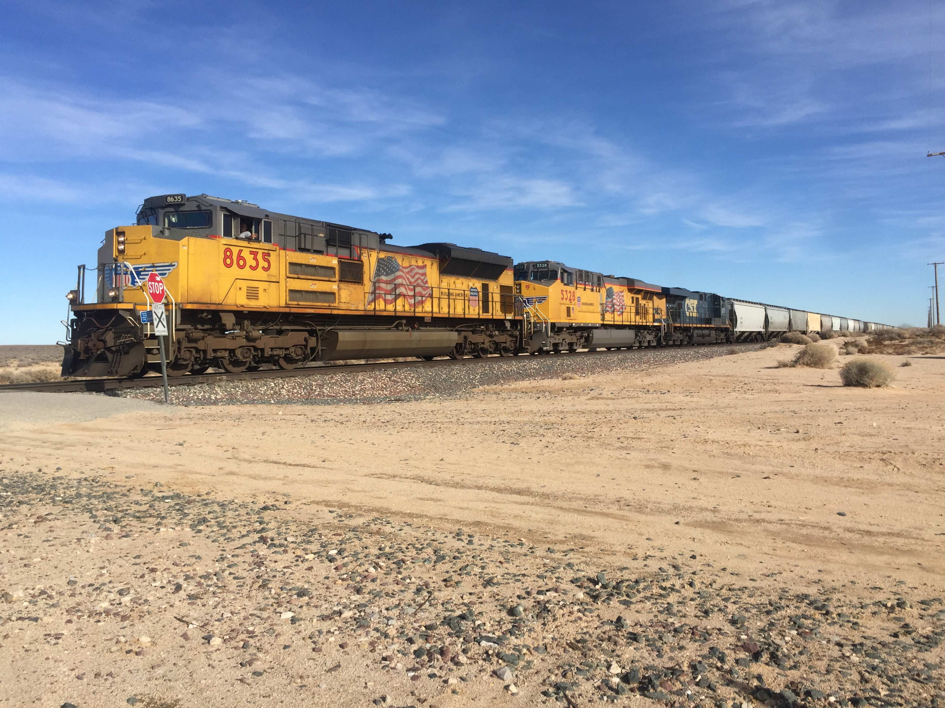 A freight train with yellow locomotives is passing through a desert landscape, with a clear blue sky overhead.