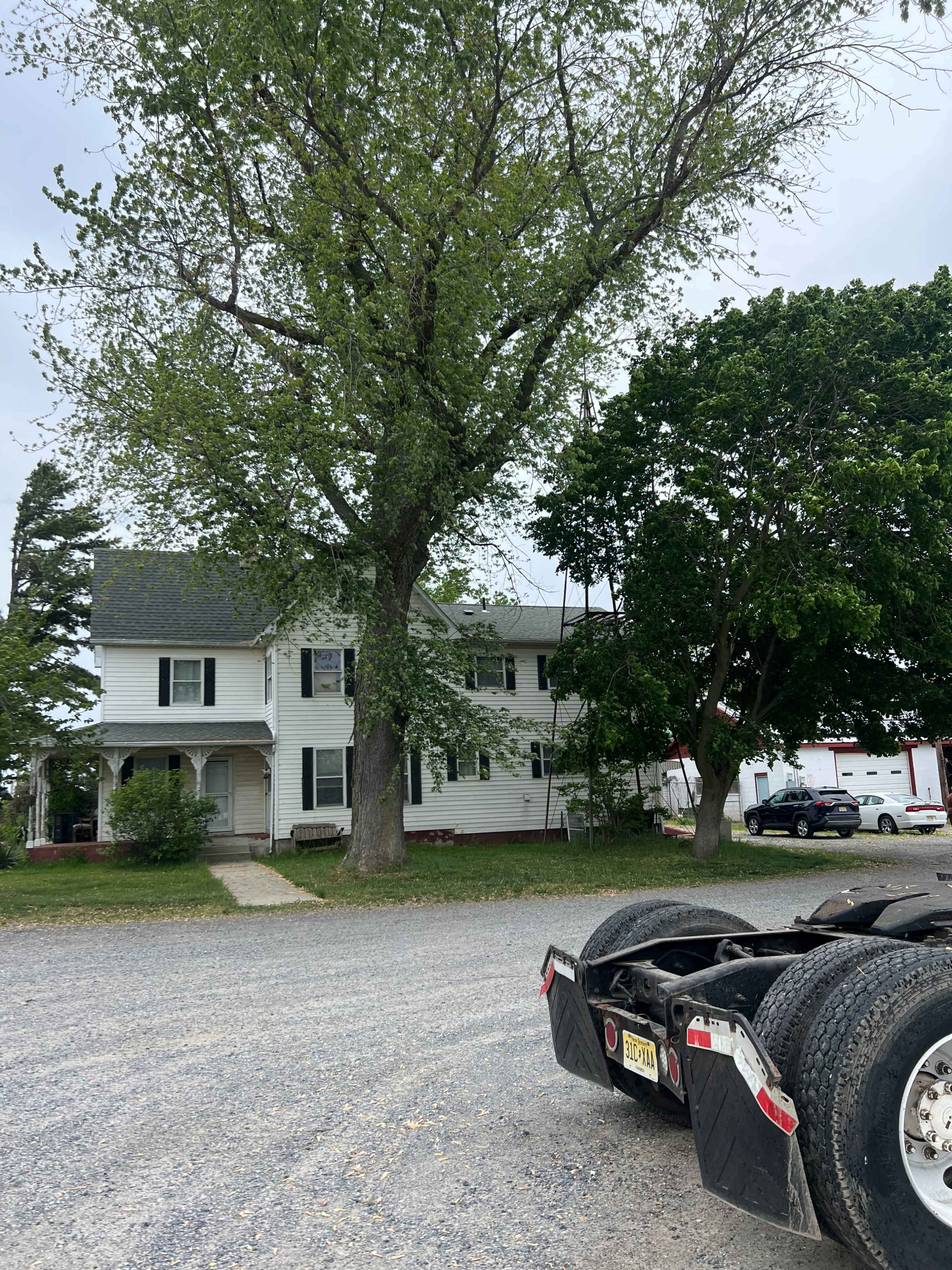 A white two-story house with a large tree in the front yard is visible next to a gravel driveway and a truck chassis parked nearby.