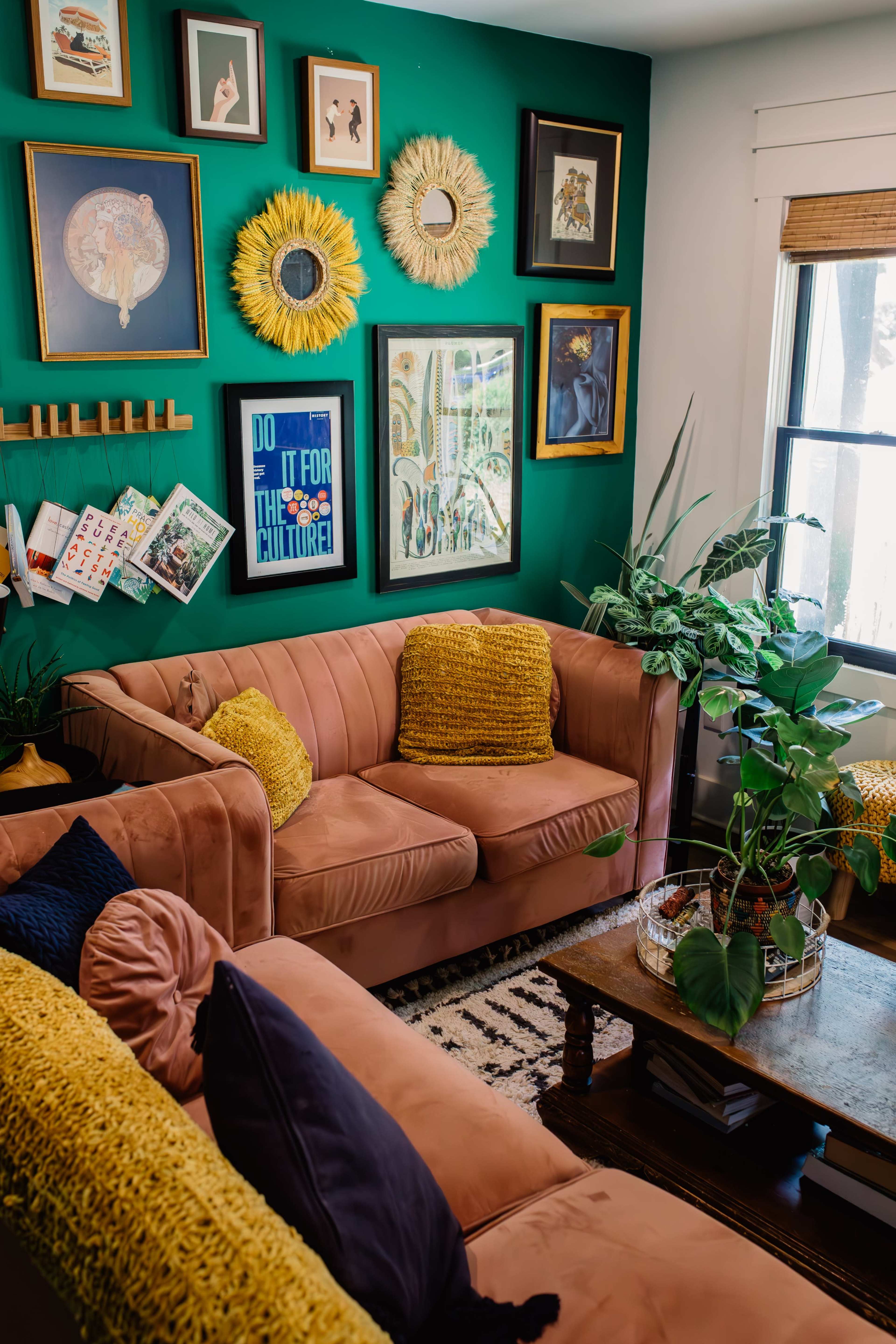 A living room features a pink sofa and a yellow-textured throw, surrounded by green walls adorned with various framed art pieces and decorative mirrors.