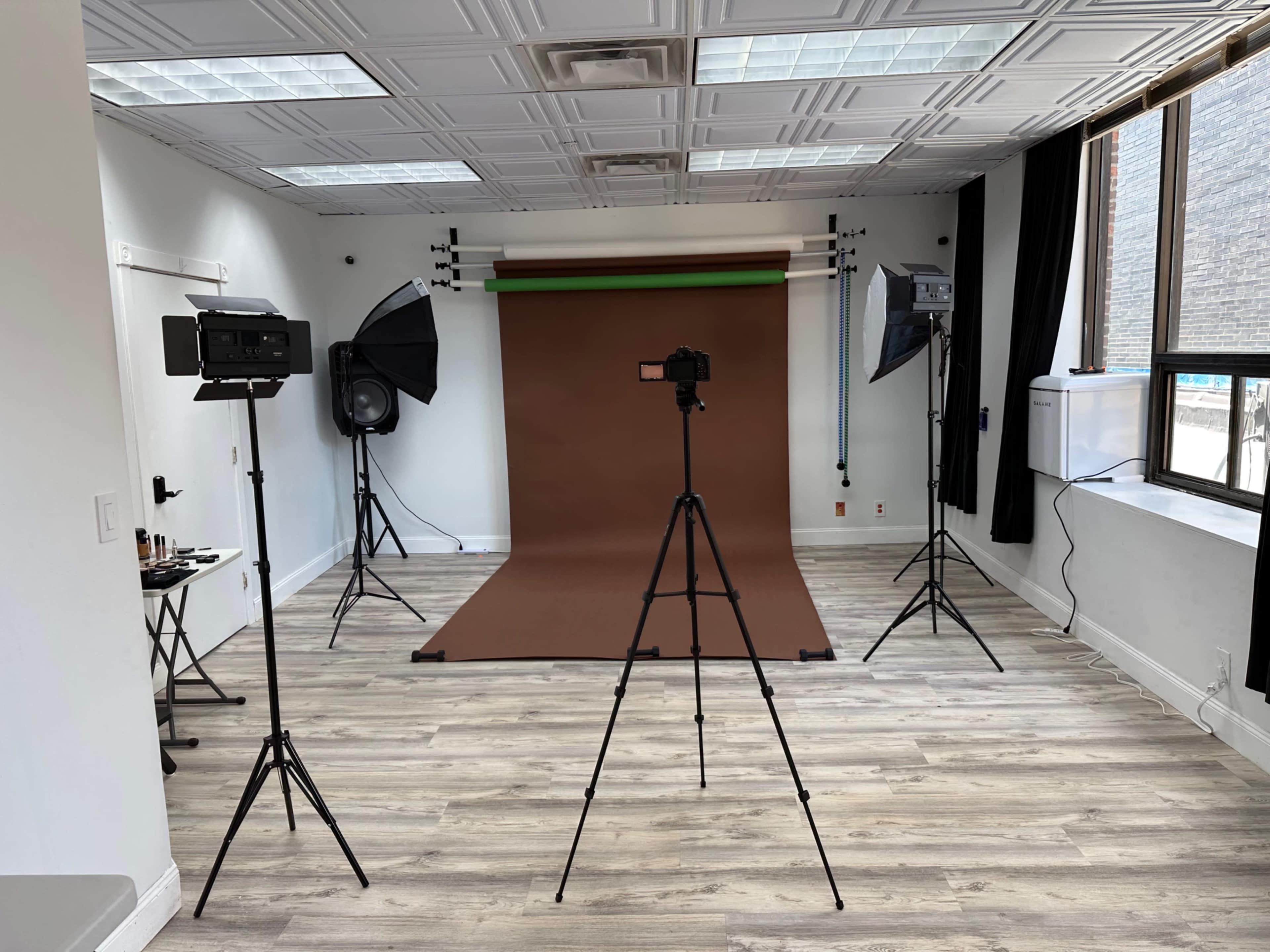 The image shows a photography studio with a brown backdrop, two studio lights, a large speaker, and tripods arranged for a photo shoot.