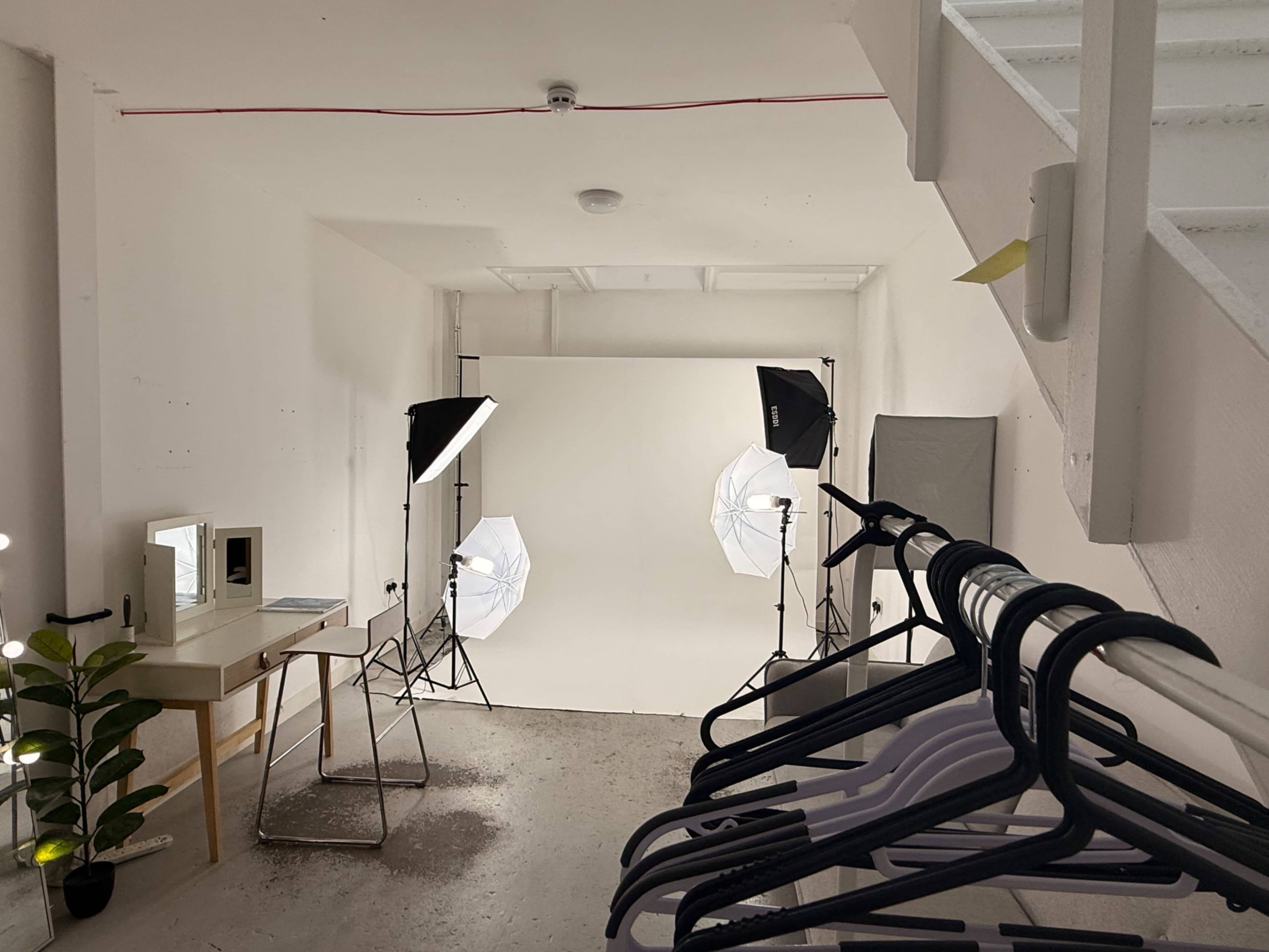 The image shows a minimalistic studio space with a white backdrop, two light sources, a dressing table with a mirror, and a rack of hangers.