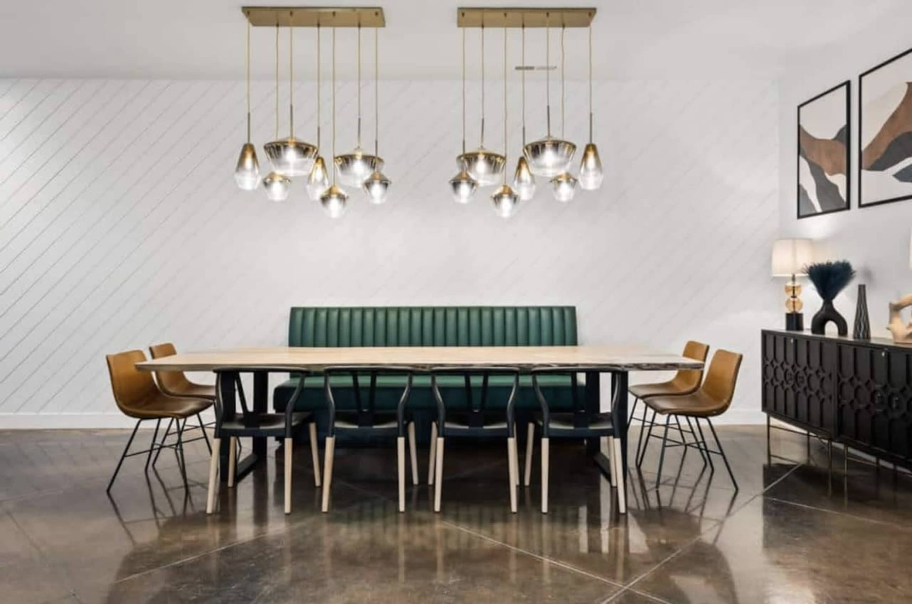 The image shows a dining area featuring a long table surrounded by chairs, with a green upholstered bench and several pendant lights hanging above.