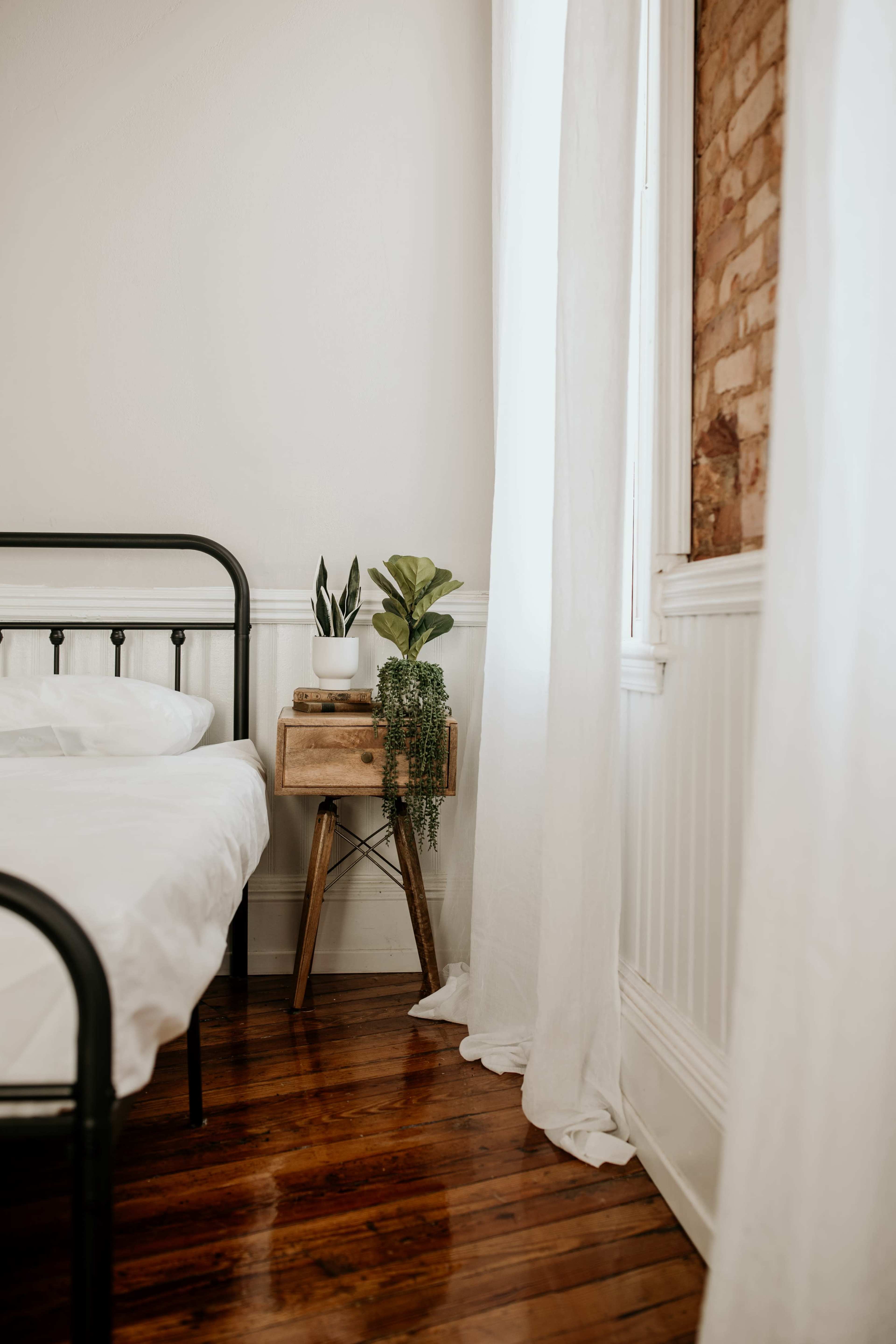 A metal bed frame is positioned next to a wooden nightstand with two potted plants, set against a light-filled room with white curtains and exposed brick wall.